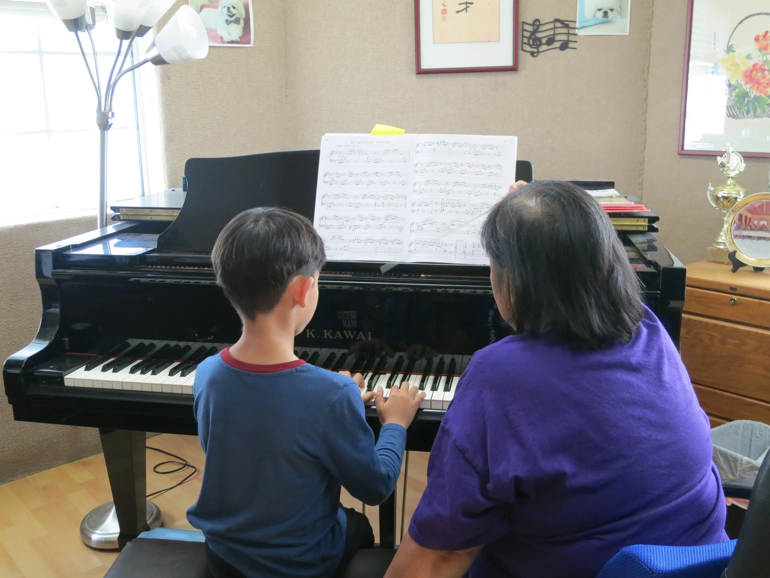 A student and a teacher are sitting at a grand piano, practicing piano together with sheet music on the stand. (The Cross Schools of Education - Walnut, CA)