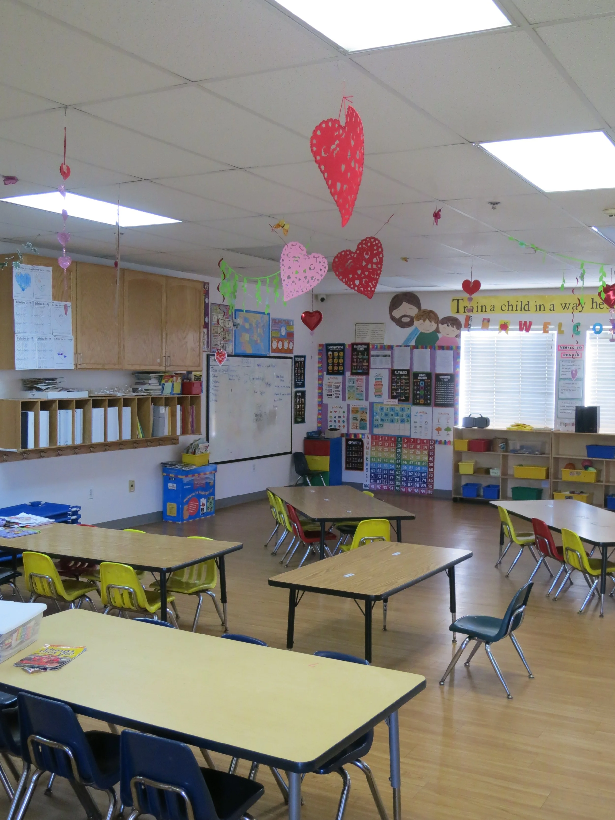 A private preschool classroom with colorful tables and chairs, decorated with hanging paper hearts and butterflies, and educational posters on the walls. (The Cross Schools of Education - Walnut, CA)
