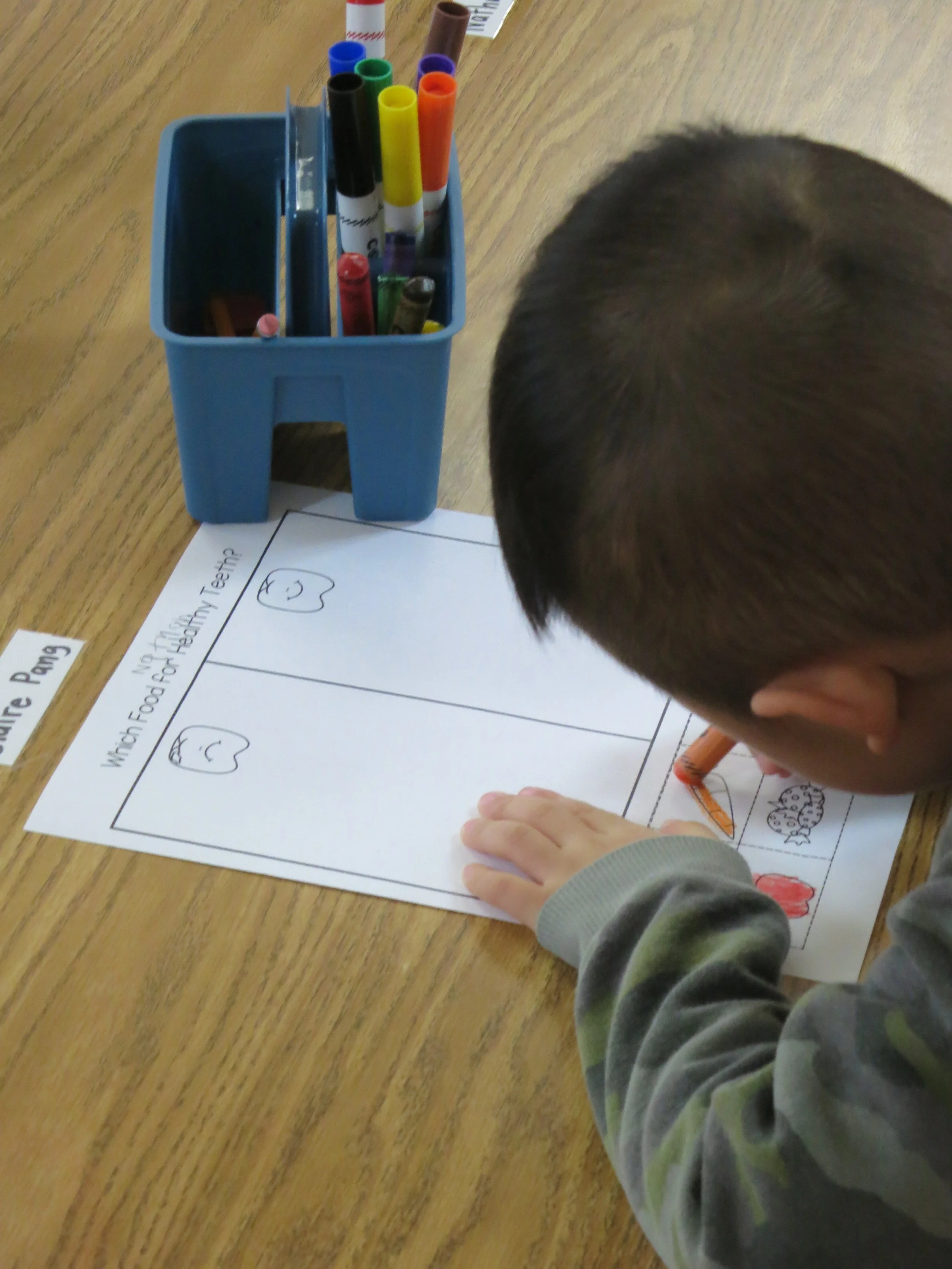 A private school student with short dark hair is coloring a worksheet about matching food to the Earth, using an orange crayon. The worksheet has hand-drawn images of a carrot, potato, and apple, and is titled 'Match Food for Healthy Earth.' There is