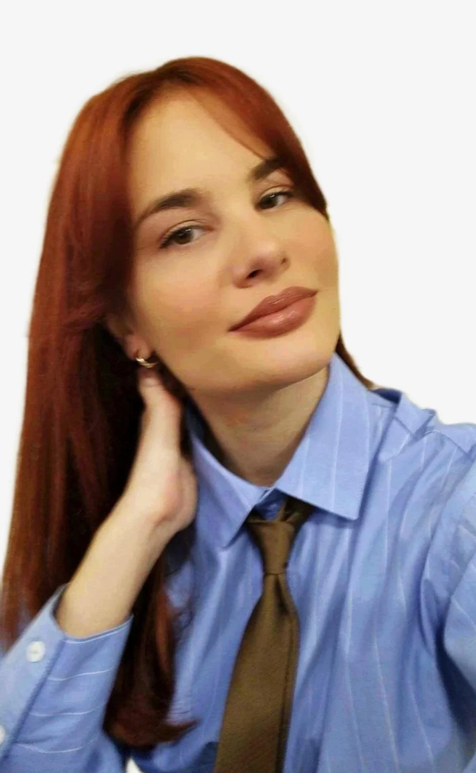 A woman with long red hair, wearing a light blue button-up shirt, a dark brown tie, and small hoop earrings, posing against a plain white background.