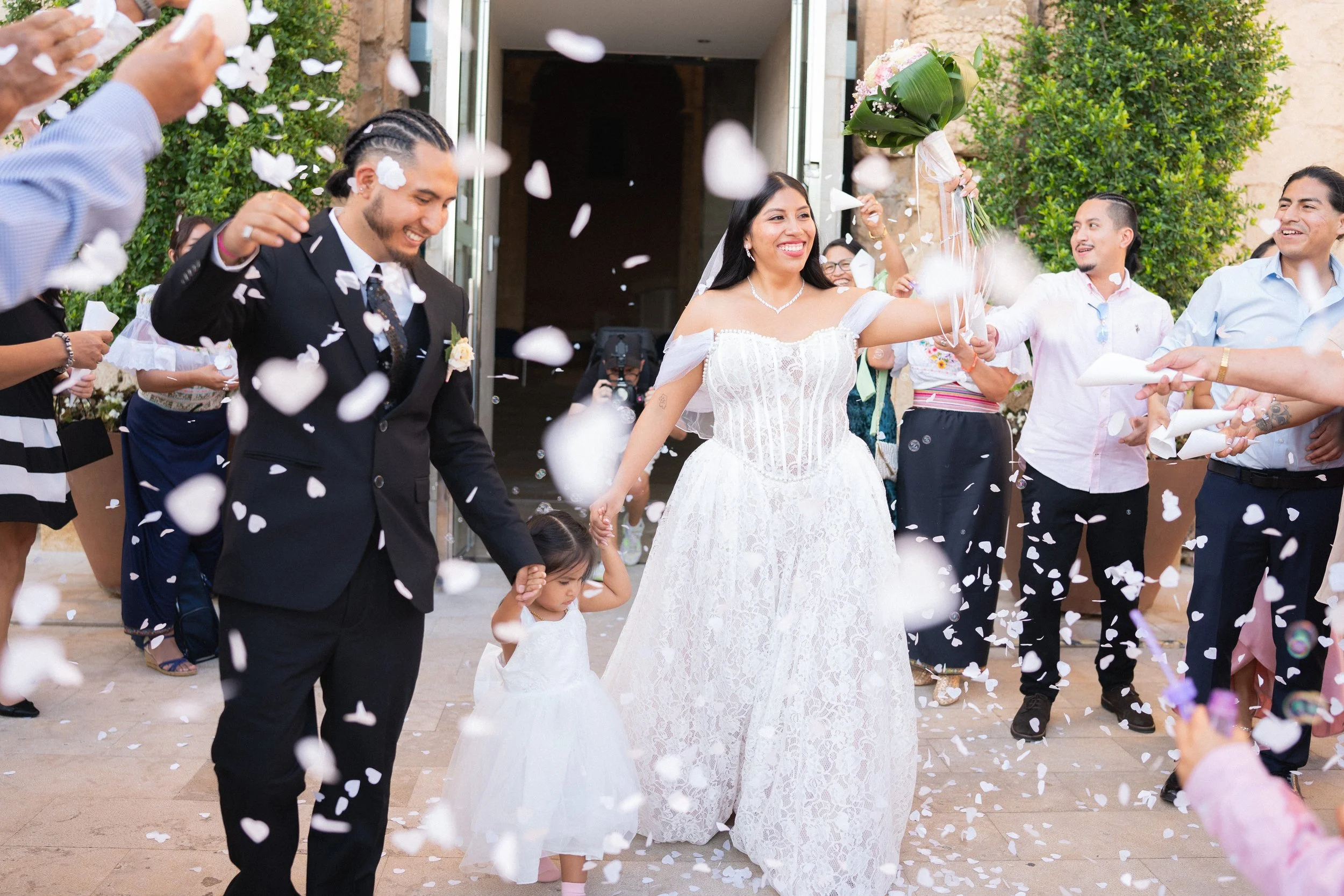 Imagen de dos personas el día de su casamiento, con lluvia de petalos de flores y su hija de la mano.