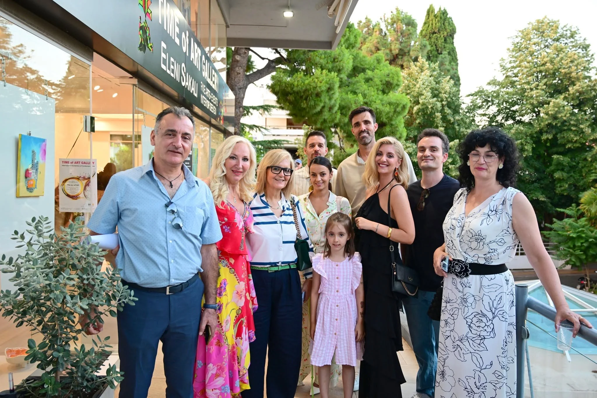 Group photo of adults and a child outside Time of Art Gallery, with artwork and greenery visible