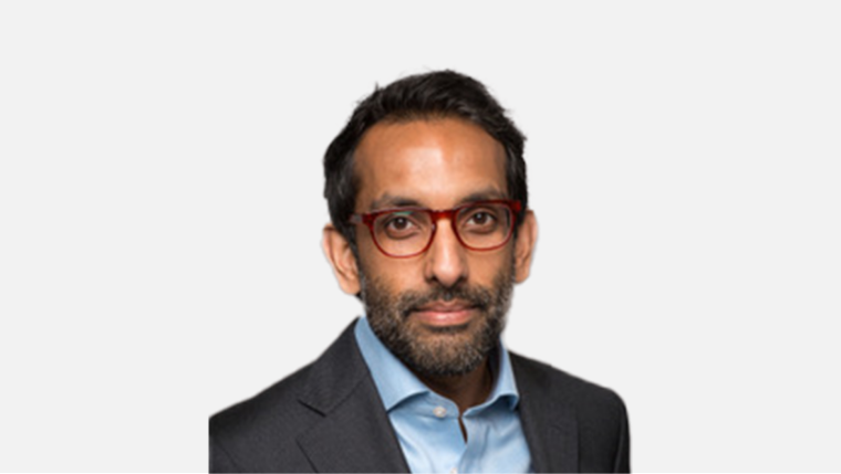 Portrait of a man with dark hair, beard, and red glasses wearing a gray suit and light blue shirt against a plain background.