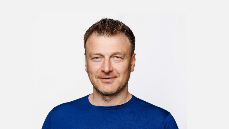 A man with short brown hair, blue eyes, and light skin, wearing a blue shirt, looking at the camera with a slight smile.