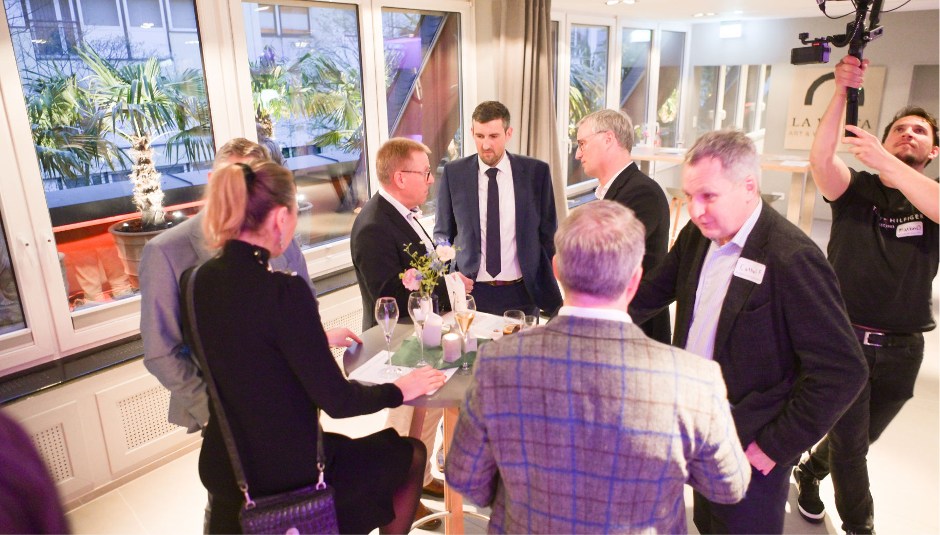 Group of people gathered around a high table at a social event, with some holding drinks, standing near large windows with a view of potted plants outside. A man on the right is operating a camera mounted on a stabilizer.