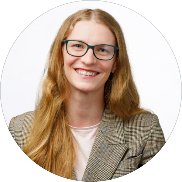 A woman with long red hair and glasses smiling, wearing a plaid blazer over a light-colored top.