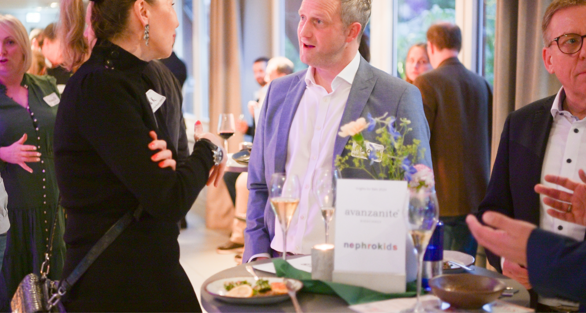 People talking at a social event with a table featuring a sign that reads "avanzite" and "nephrokids," glasses of wine, and a plate of food.