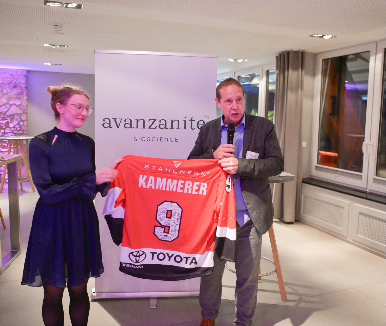 A man holding a microphone and a woman holding a sports jersey in a decorated indoor setting, with a banner behind them reading 'avanzanite BIOSCIENCE'.