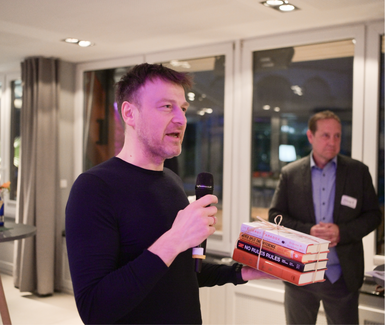 Man speaking into a microphone holding a stack of books in a room with large windows and curtains.