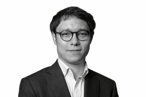 Black and white portrait of a man wearing glasses, a suit, and a collared shirt, looking at the camera.