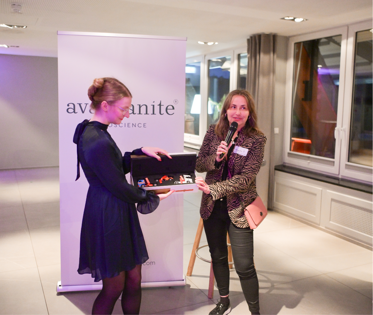 A woman in a black dress holding a box of colorful watches, while another woman with a microphone, wearing a zebra-striped blazer, stands beside her at an event, with a backdrop displaying the logo 'avatonite'.