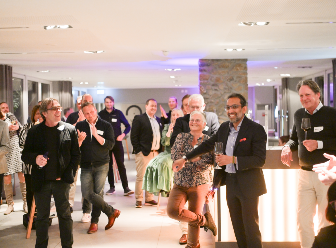 Group of people at a social event, some holding wine glasses, smiling and talking indoors with a bar area and a stone pillar in the background.