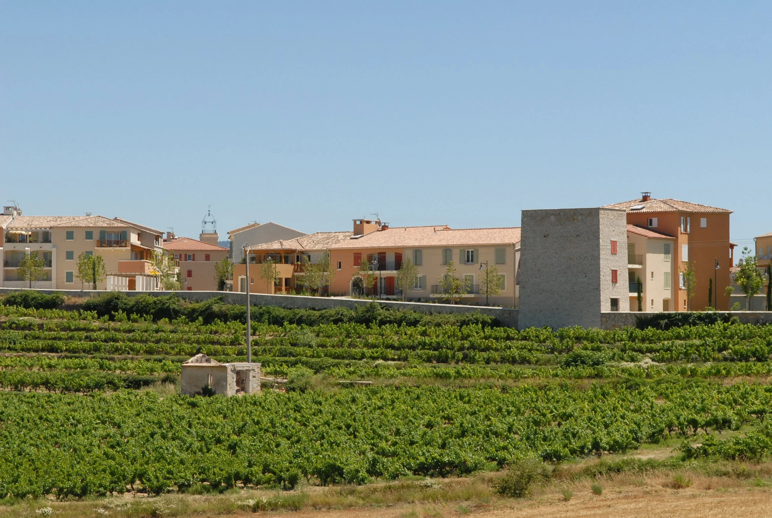 Vignoble avec de petites maisons colorées sous un ciel bleu, quelques arbres et un bâtiment en pierre au premier plan.