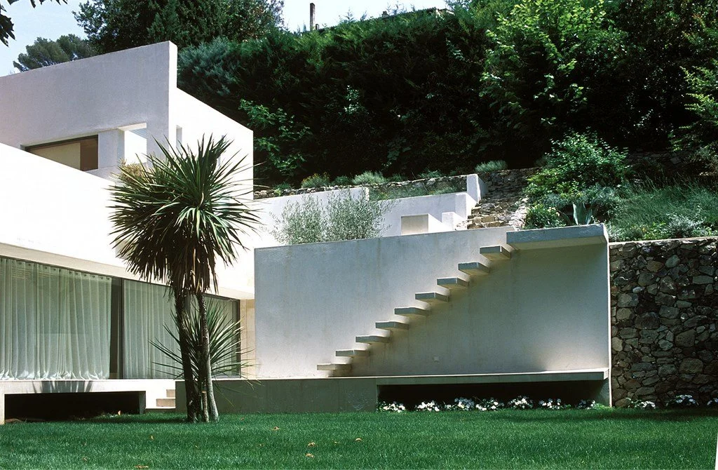 Maison moderne blanche avec escalier extérieur, grand fenestrage avec rideaux, arbre en premier plan, terrasse surélevée, jardin bien entretenu, sur une pente avec végétation dense au fond.
