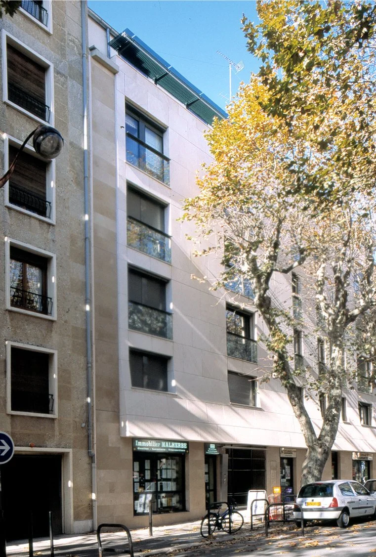 Bâtiment résidentiel moderne avec des fenêtres et un magasin immobilier au rez-de-chaussée, arbres avec des feuilles jaunes, une voiture blanche et un vélo garés devant, en plein jour.