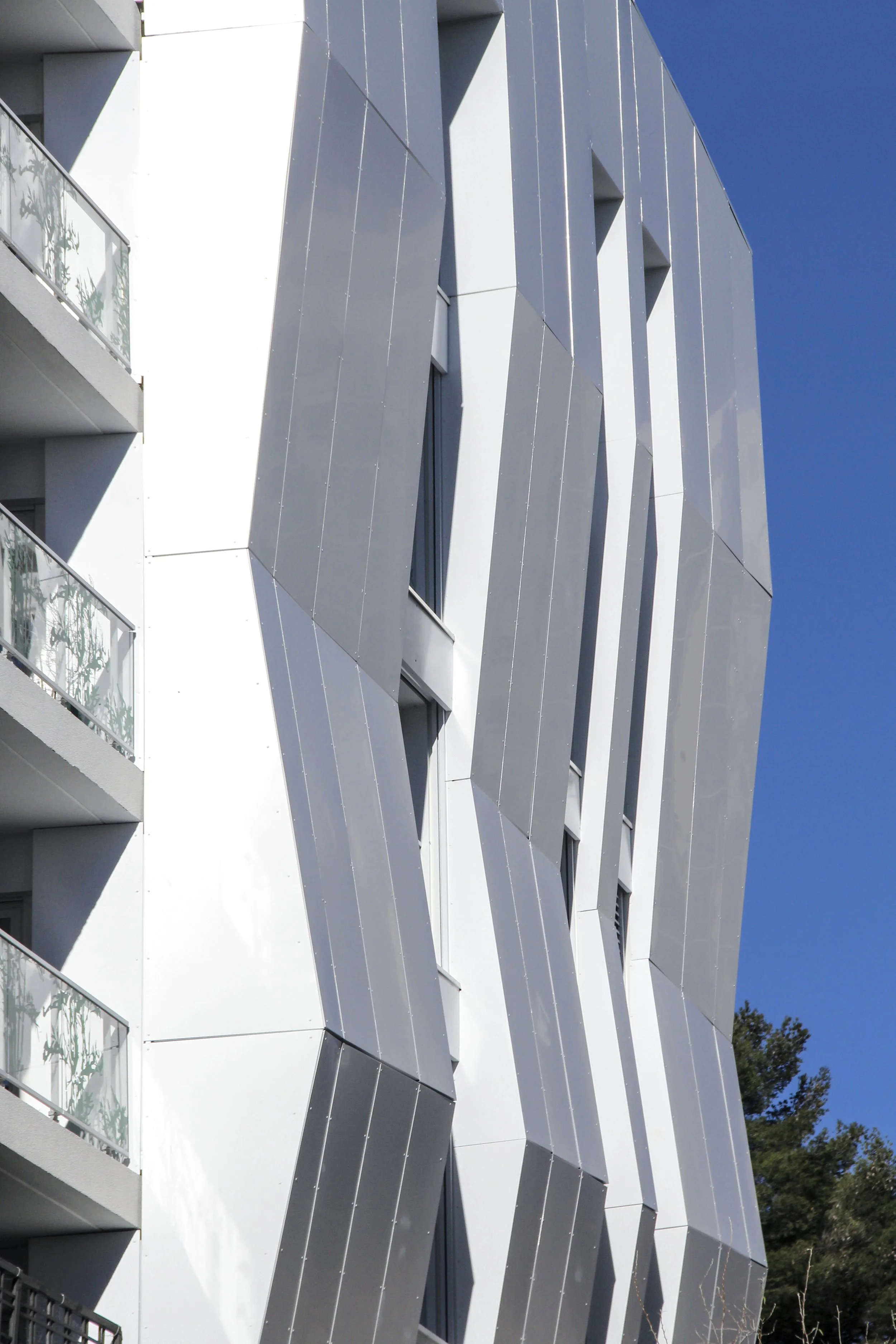 Façade moderne d'un bâtiment avec des panneaux métalliques gris en relief et des fenêtres rectangulaires, avec des balustrades en verre sur le côté gauche, sous un ciel bleu clair.
