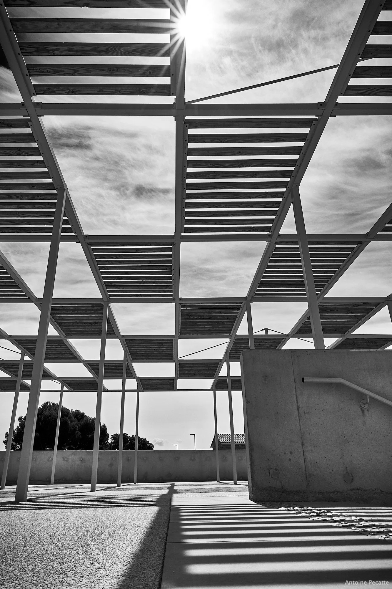 Structure en acier et bois en plein air avec des lattes de bois formant un plafond partiellement ouvert, ombrageau sol, ciel nuageux en arrière-plan, photo en noir et blanc.
