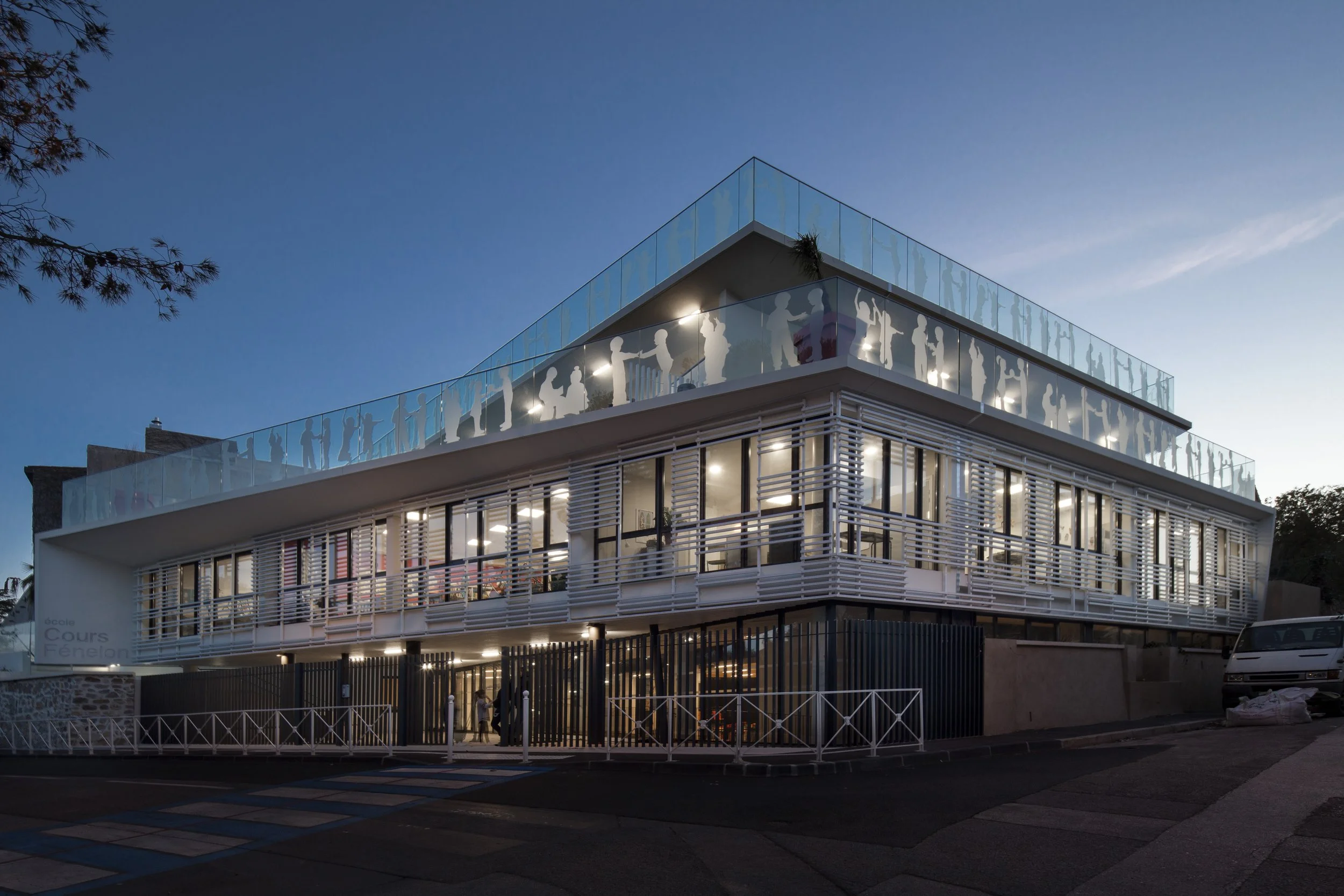 Bâtiment moderne avec plusieurs étages, illuminé en soirée, avec des silhouettes de personnes sur le balcon supérieur en verre.