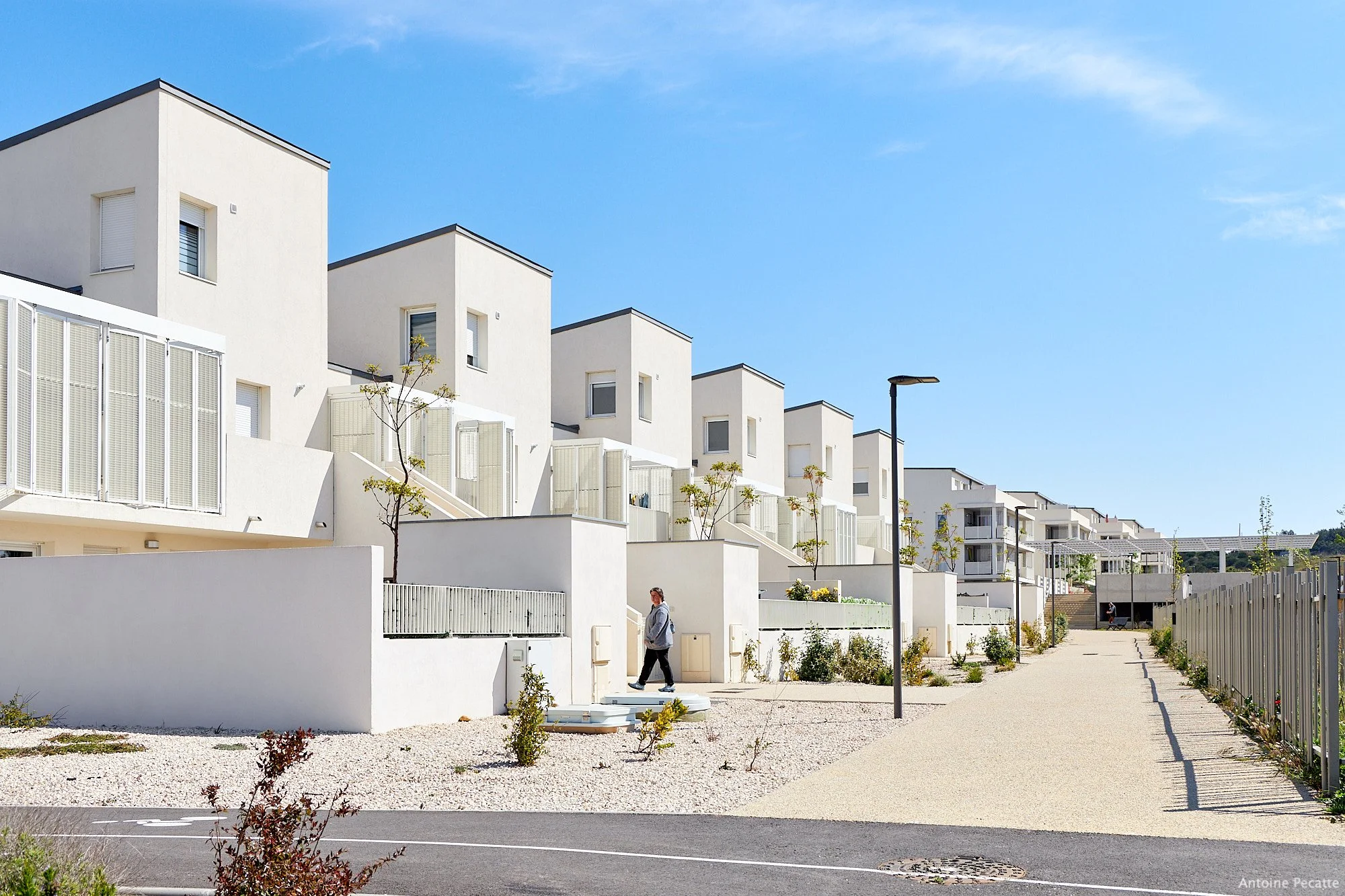Immeubles résidentiels modernes blancs avec un ciel bleu clair, trottoir et lampadaires, quelques arbres et une personne marchant