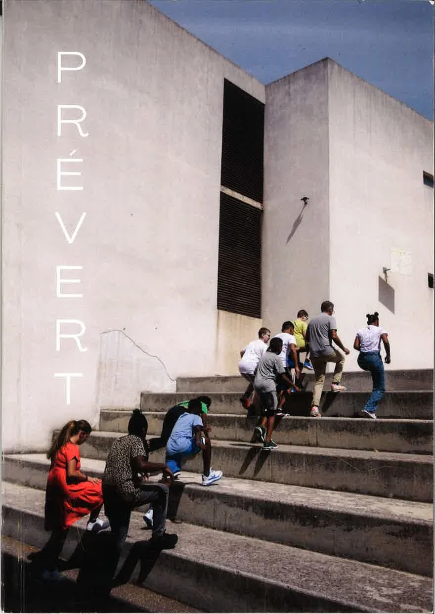 Une groupe de personnes, principalement des enfants, en train de monter des escaliers en pierre devant un grand bâtiment blanc moderne avec peu de fenêtres, sous un ciel bleu.