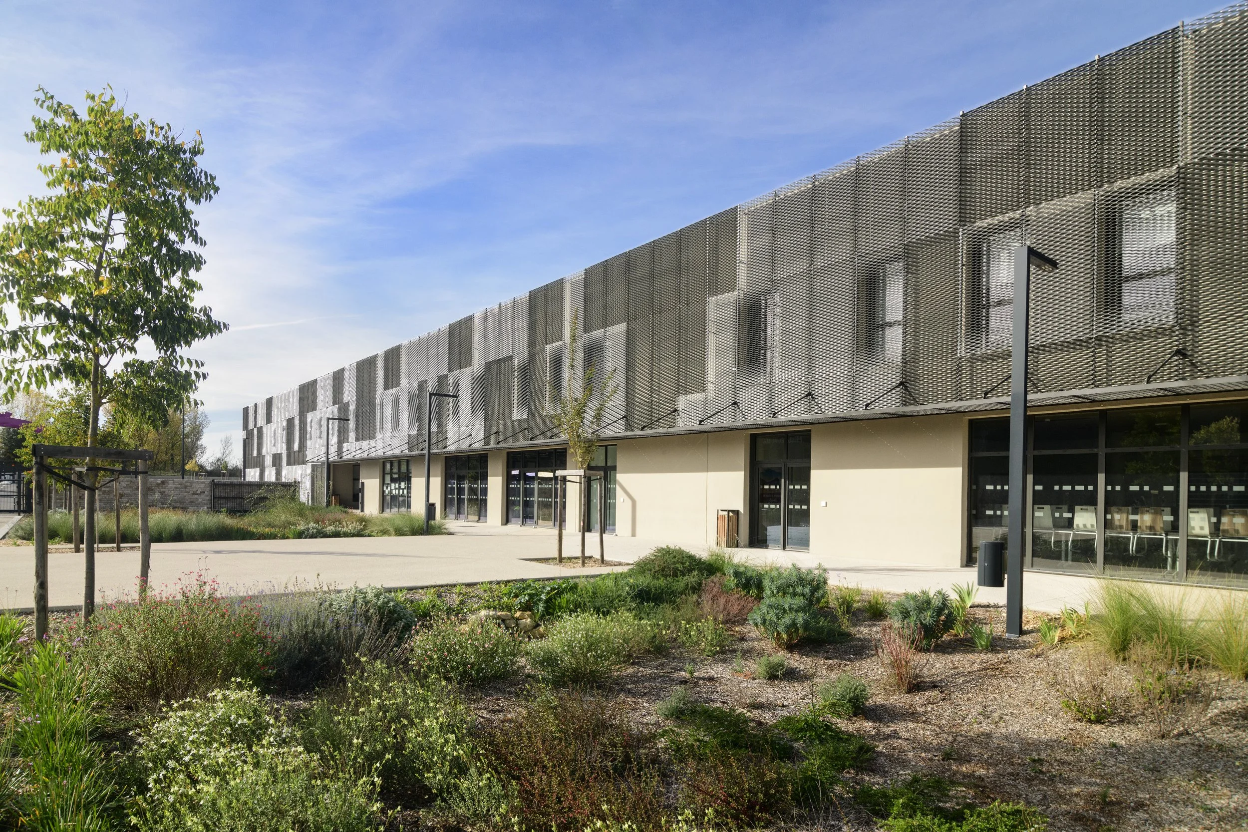 Bâtiment moderne avec une façade en métal perforé, fenêtres en verre, et un espace extérieur paysager avec des arbres, des arbustes, un lampadaire, et un ciel bleu.
