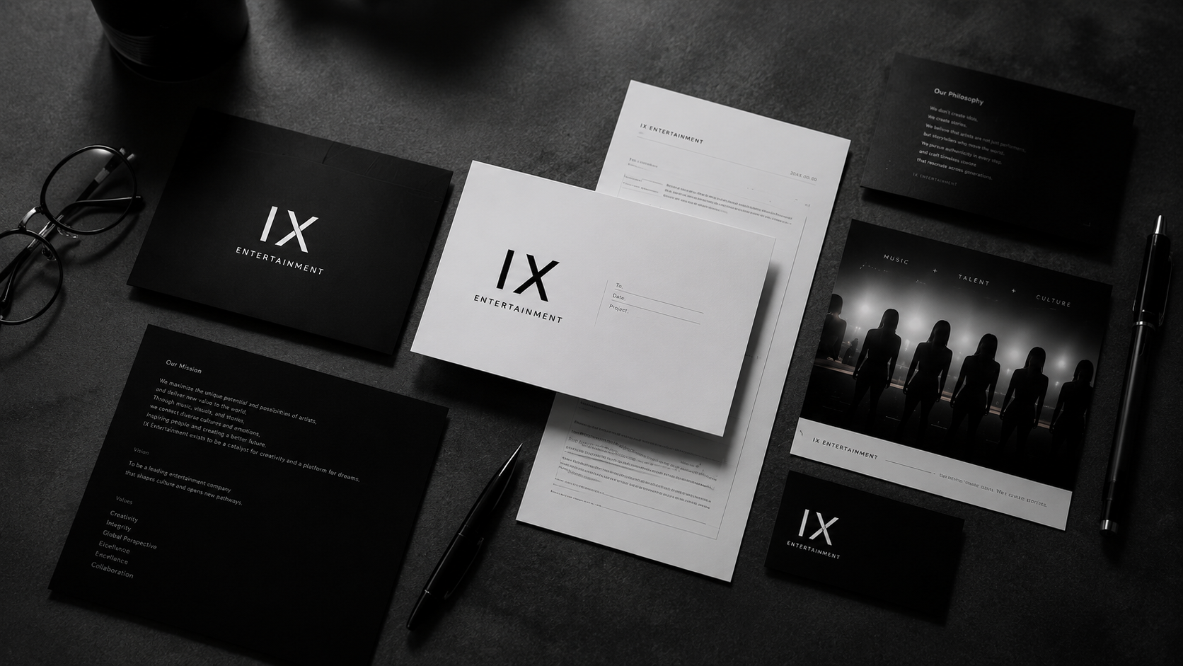 黒い背景のデスクに白と黒の書類、名刺、ペン、眼鏡が配置されている。書類には『IX ENTERTAINMENT』のロゴが見える。