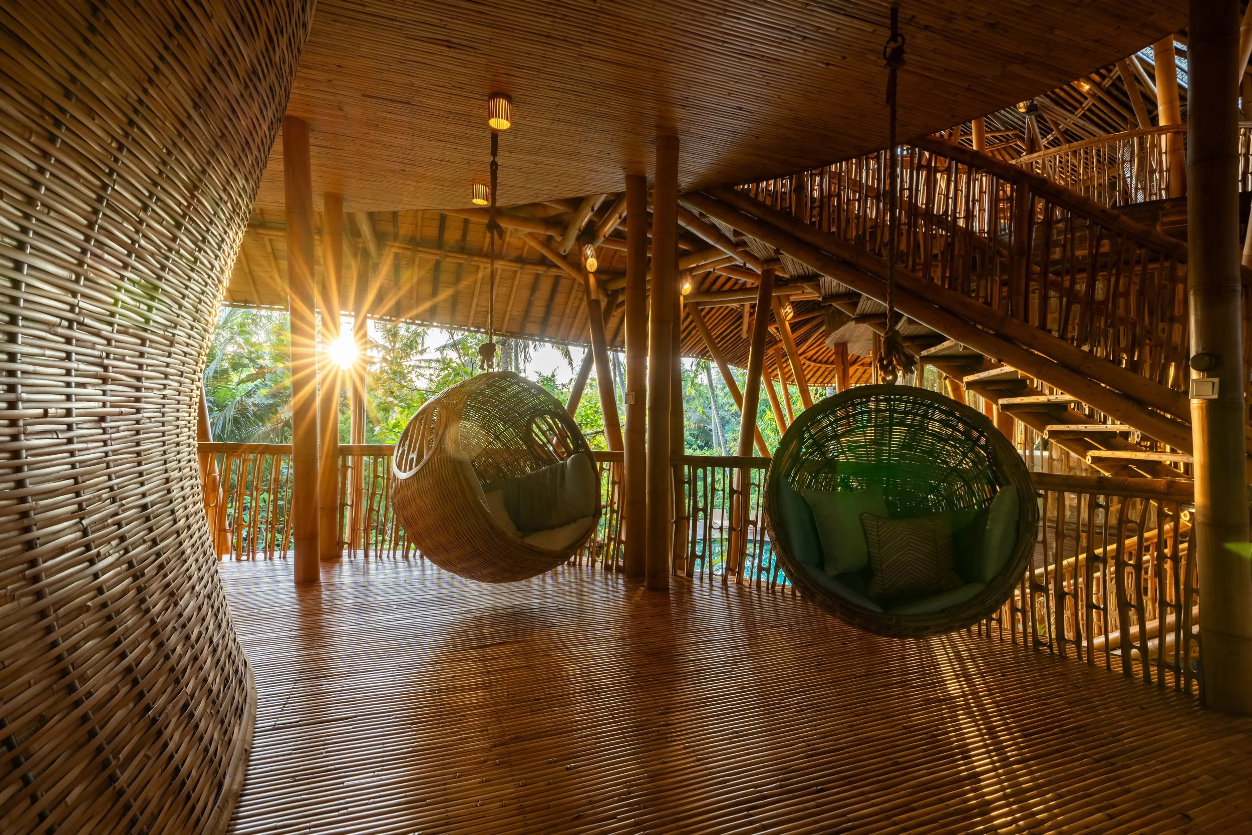 Interior of a bamboo treehouse with hanging wicker chairs, wooden staircase, and sunset view