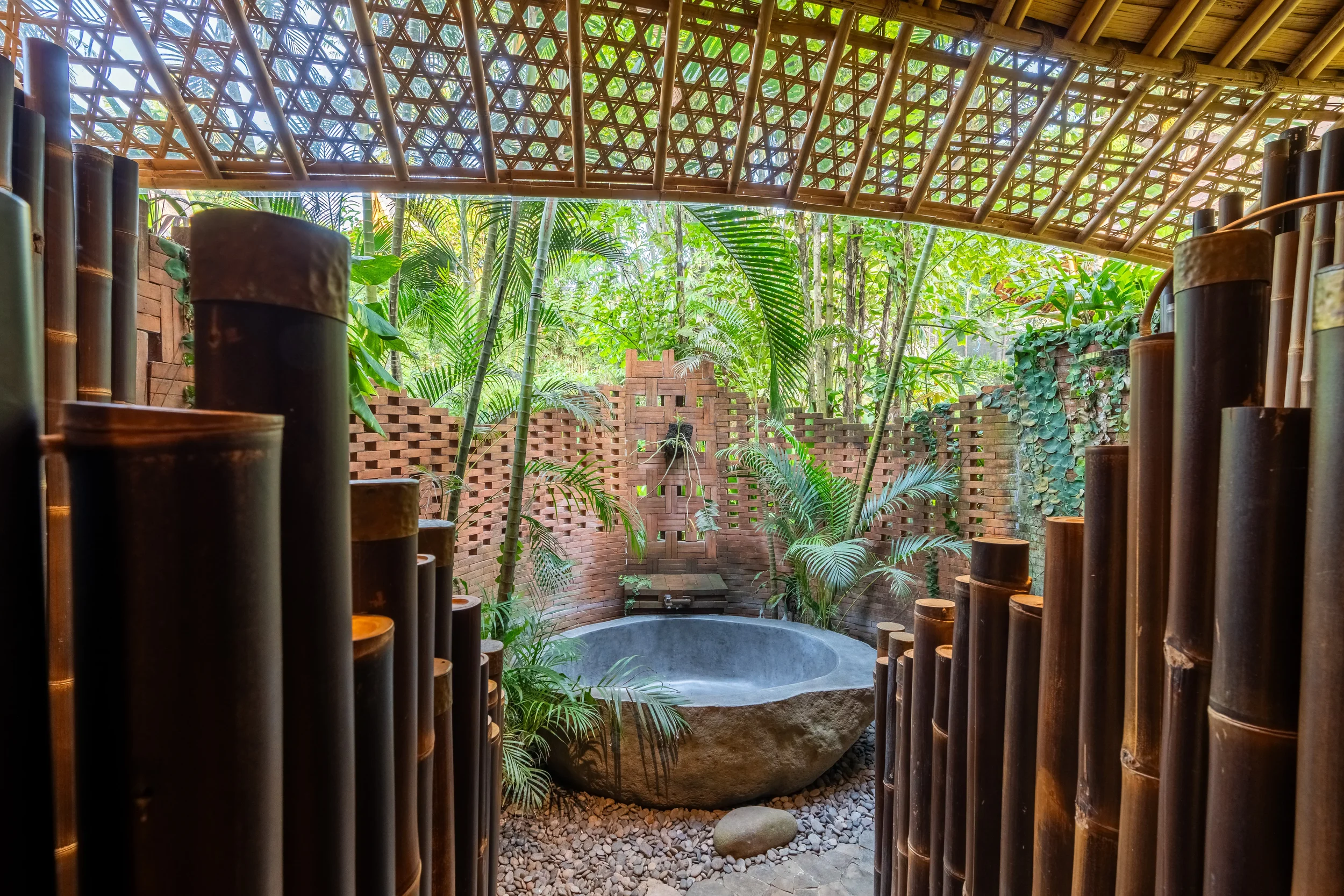 A tropical outdoor shower area with bamboo walls and a bamboo roof, featuring a large stone bowl fountain surrounded by lush green plants and trees.