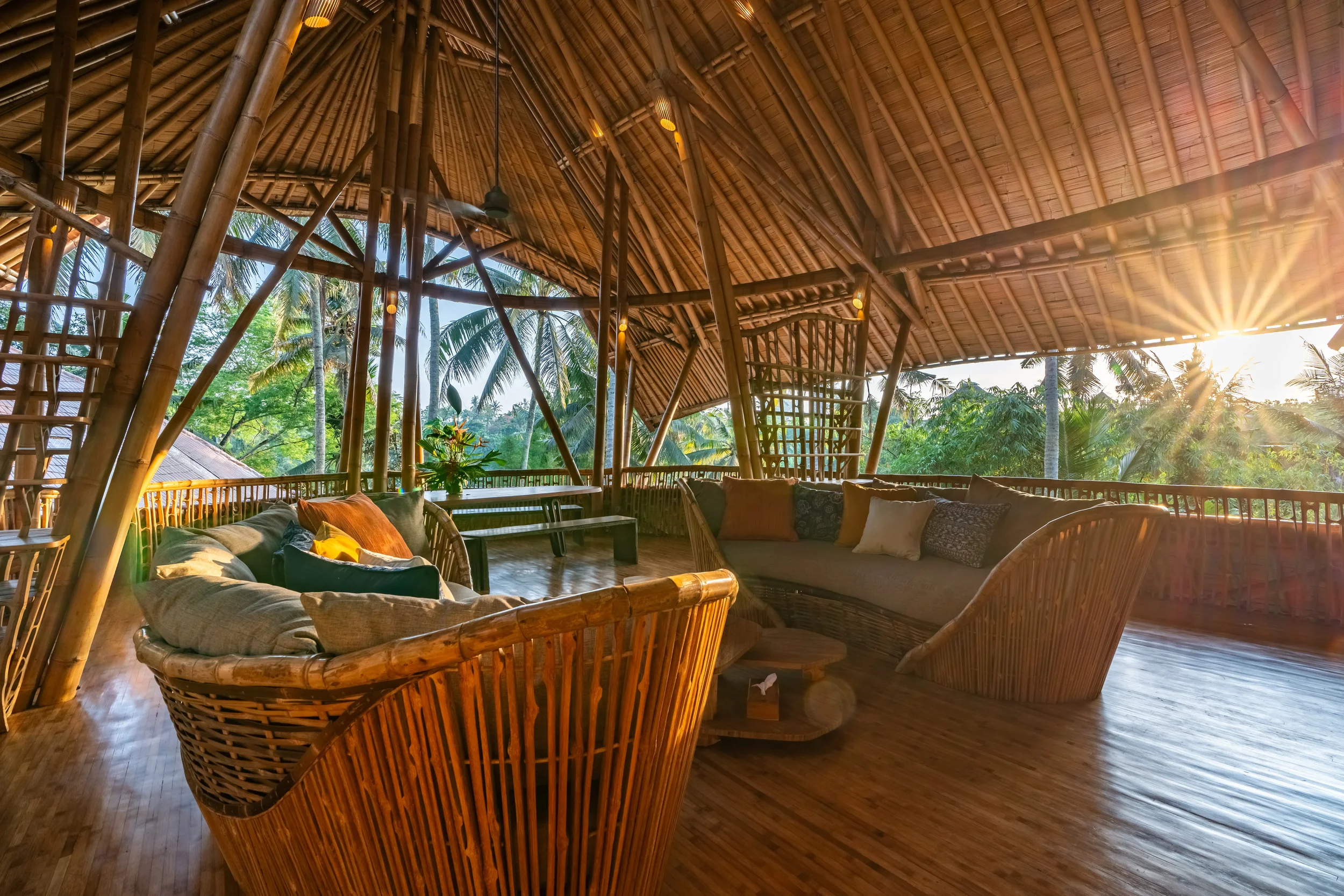 Interior of a bamboo hut with wicker furniture, including sofas and chairs, surrounded by lush green trees with sunlight filtering through.