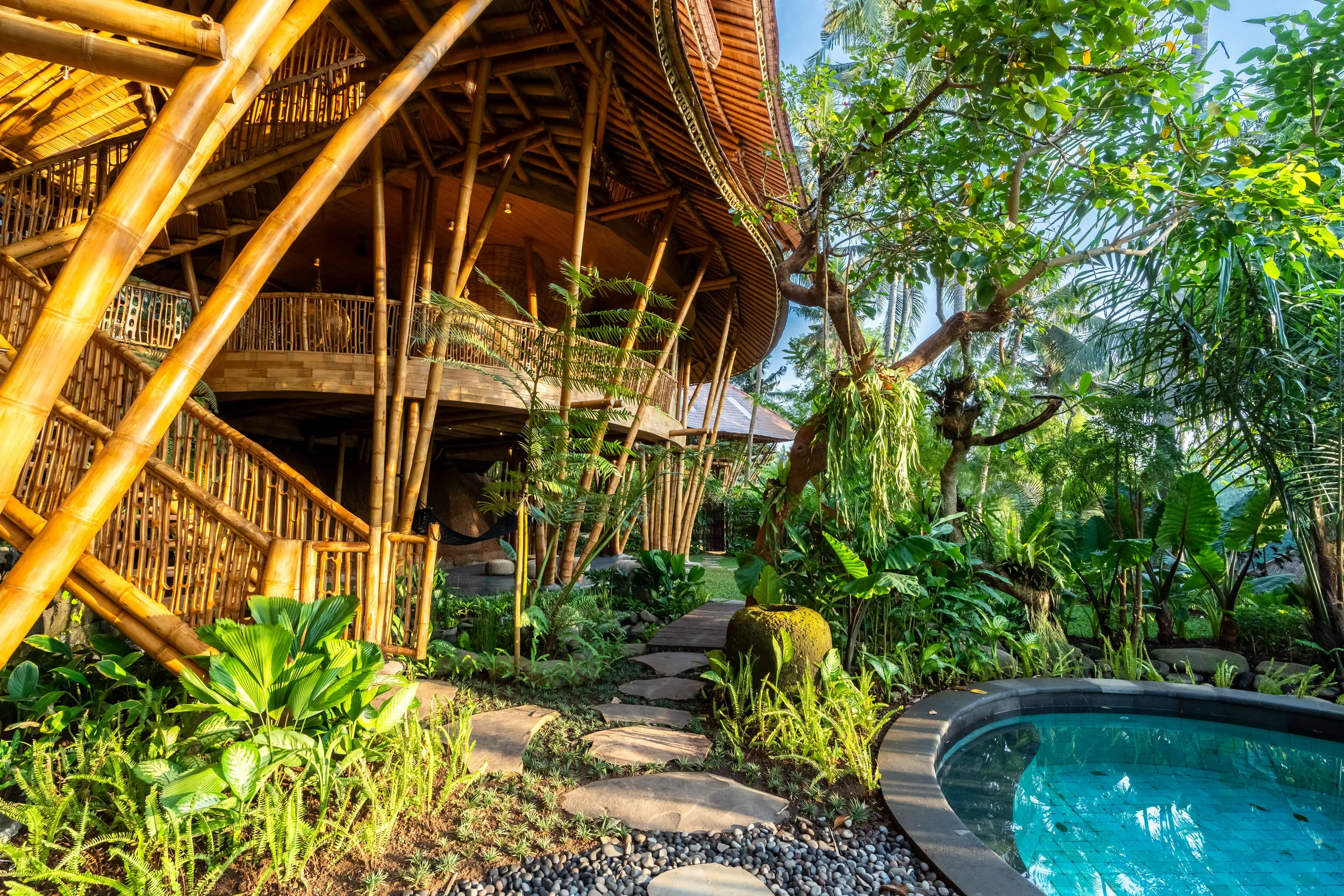Lush tropical garden with a bamboo house, pathway, greenery, and a small pool.