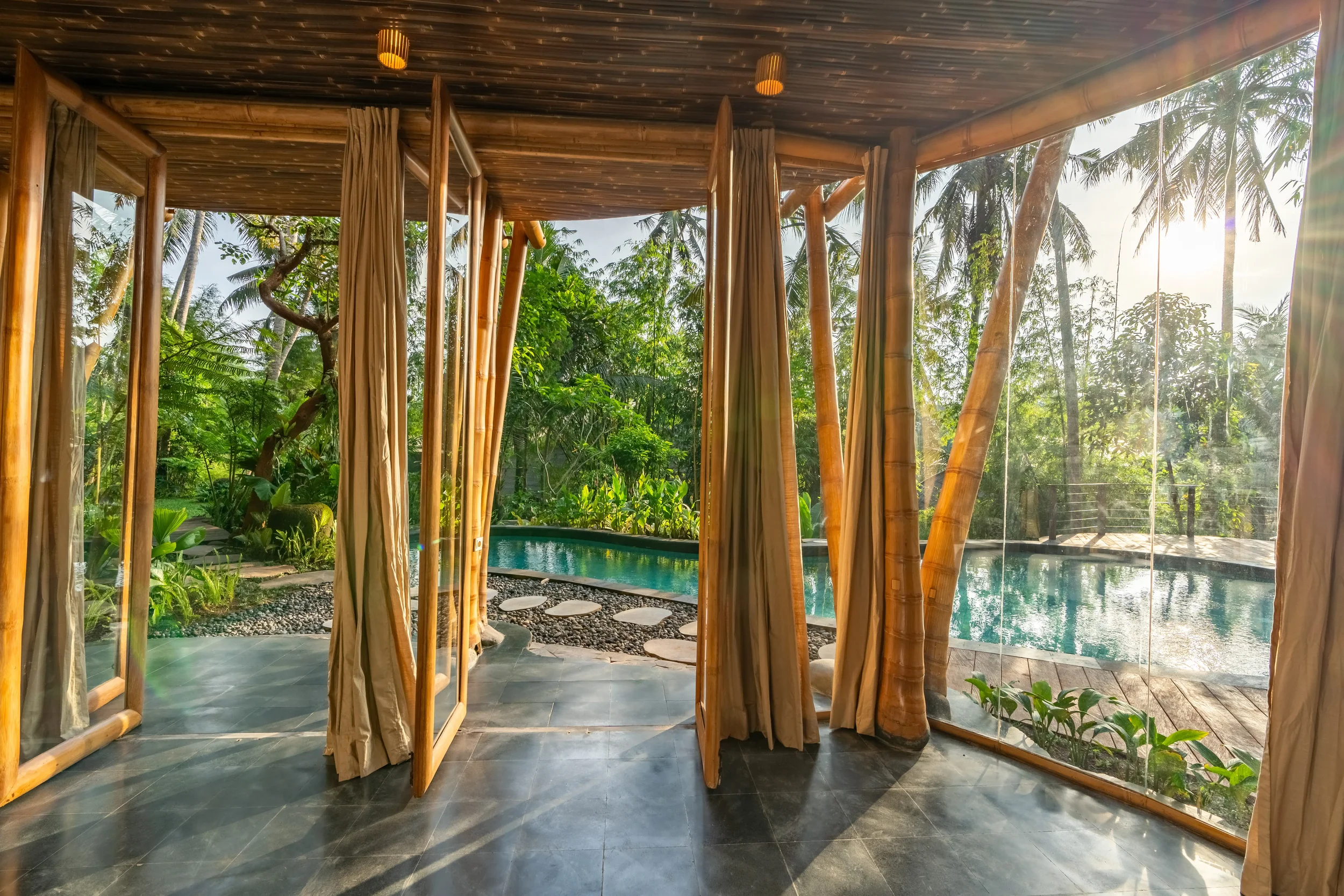 View from inside a room looking out to a lush tropical garden with a swimming pool, framed by bamboo and glass walls, with sunlight shining through trees.