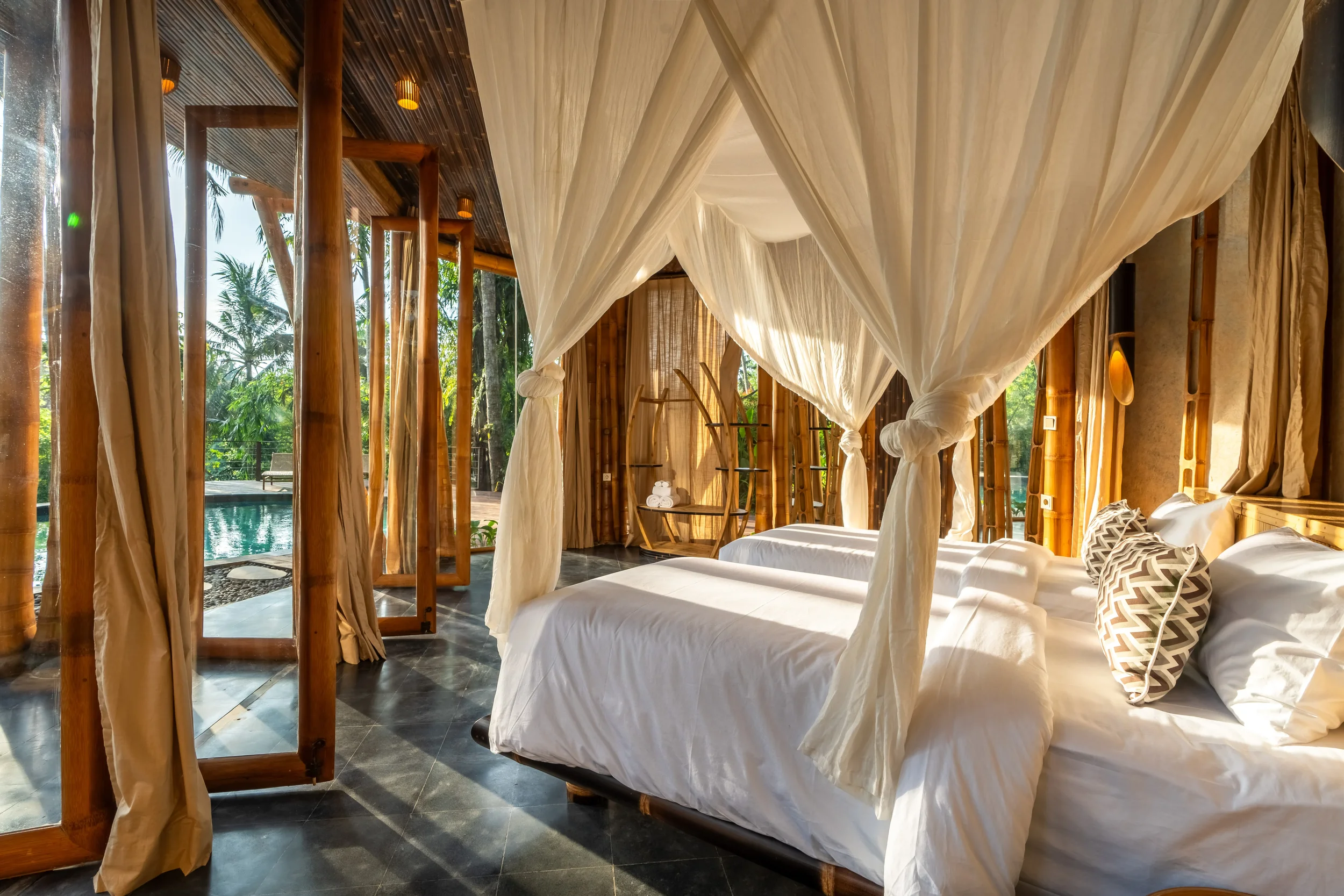 A bedroom with two beds covered with white linens and bamboo accents, surrounded by tropical greenery seen through large open windows.
