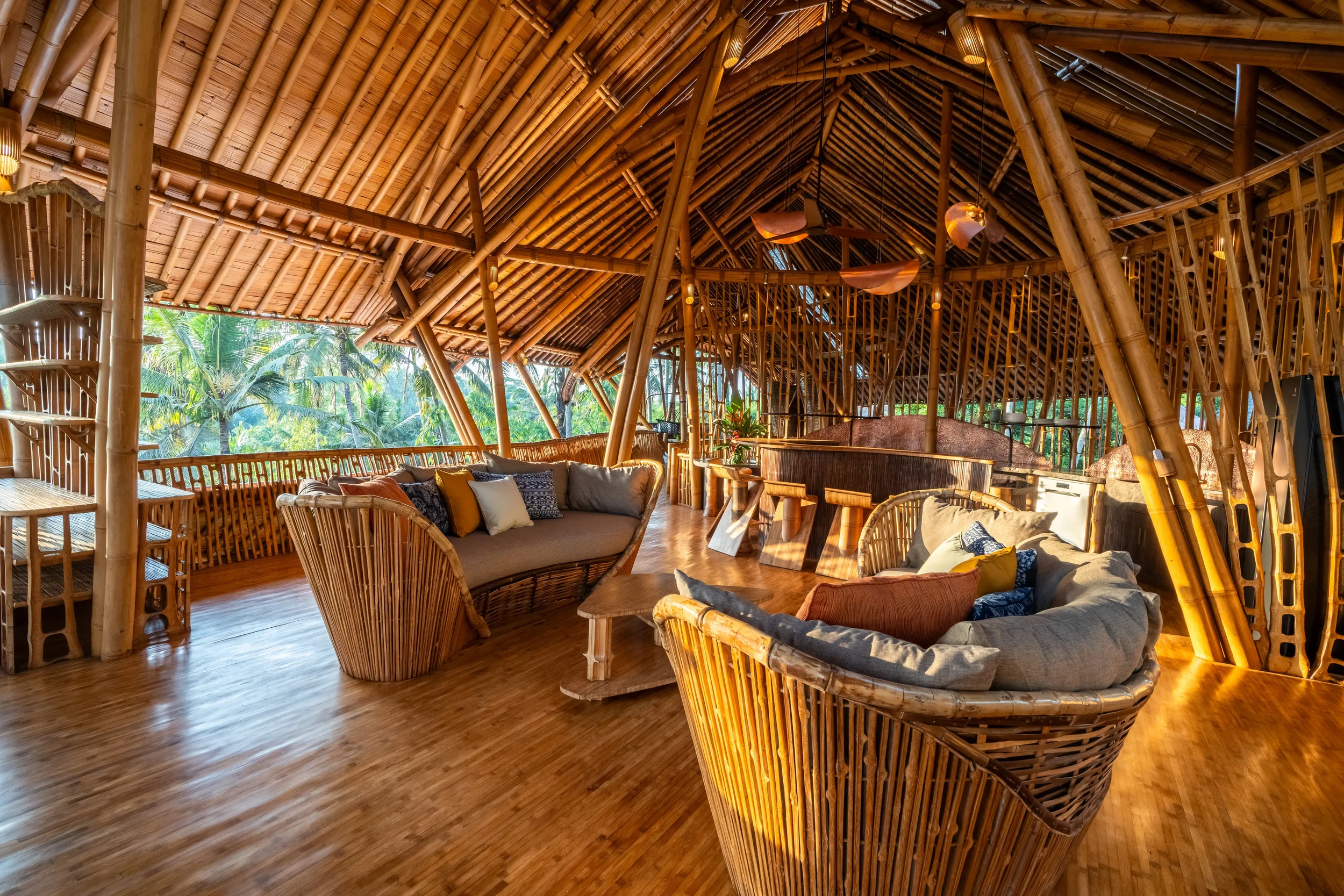 Interior of a bamboo house with woven bamboo furniture, pillows, and lush green tropical trees visible outside.