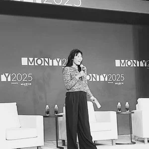 A woman speaking into a microphone on a stage with a backdrop reading 'Monty 2025 March' and two white chairs with water bottles on a table.