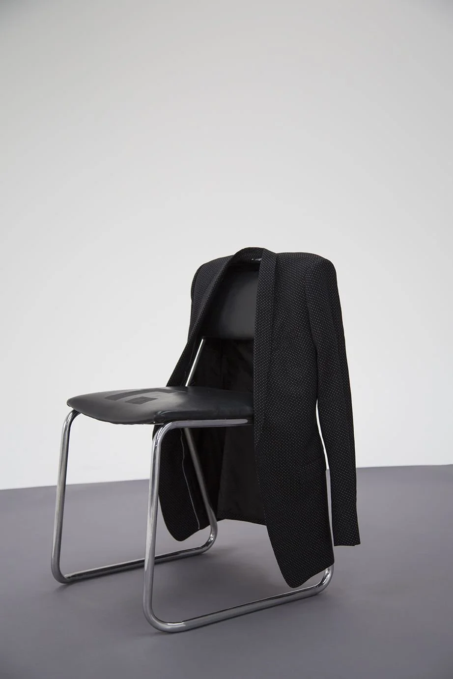 A black office chair with a black jacket draped over its backrest, set against a plain light gray wall and gray floor.
