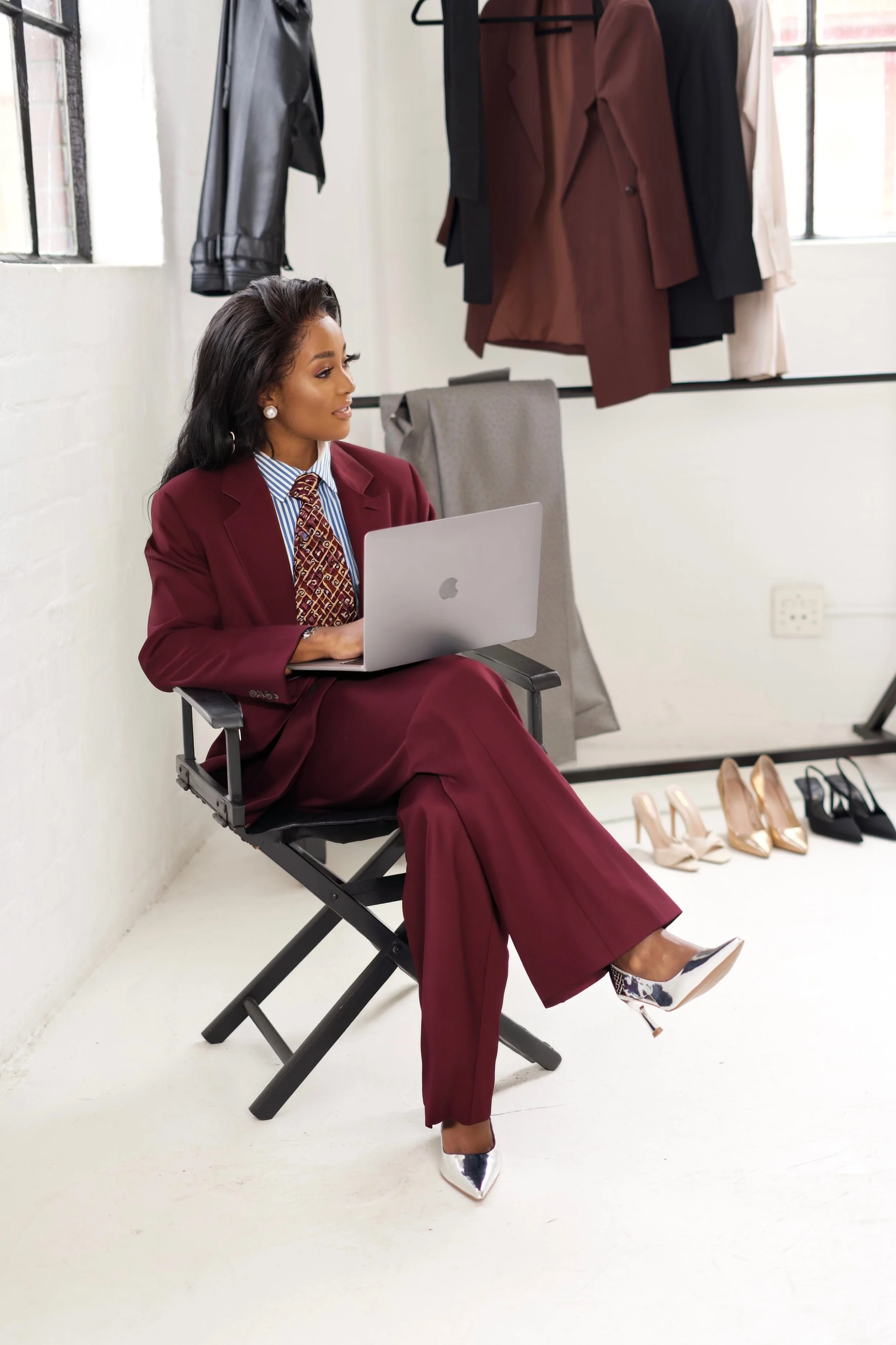 A woman sitting on a director's chair in a fashion studio, wearing a maroon suit, working on a laptop, with shoes and clothing hanging in the background.