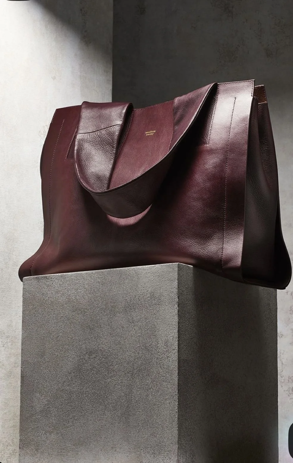 A large, dark red leather handbag on a concrete pedestal against a plain wall.