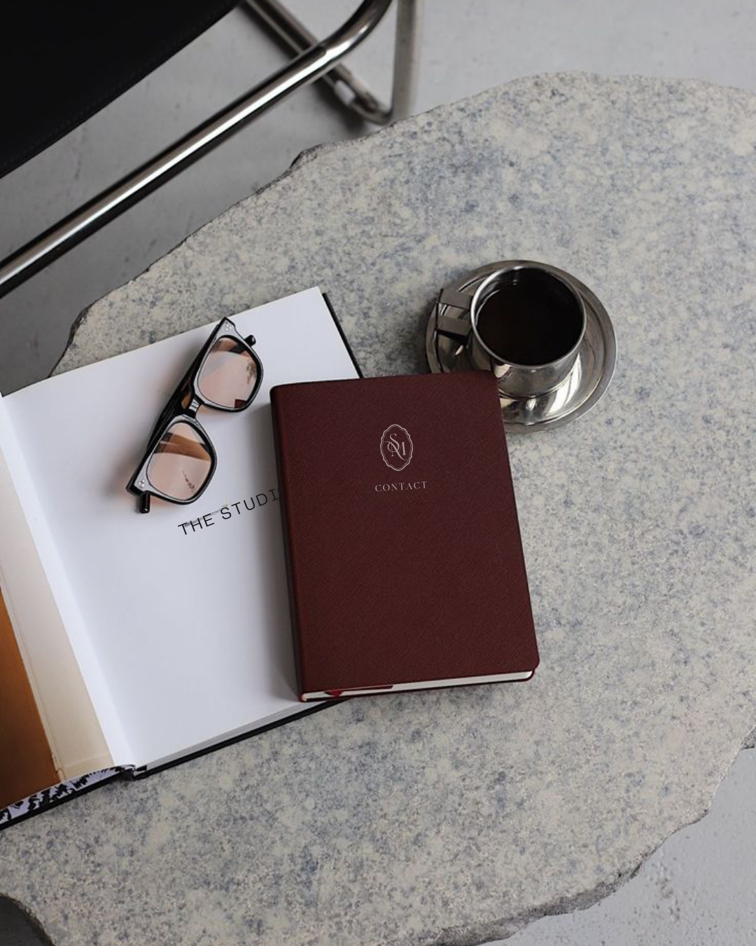 A gray stone table with a pair of eyeglasses, an open notebook with "THE STUDI" visible, a red contact book, and a cup of coffee on a saucer.