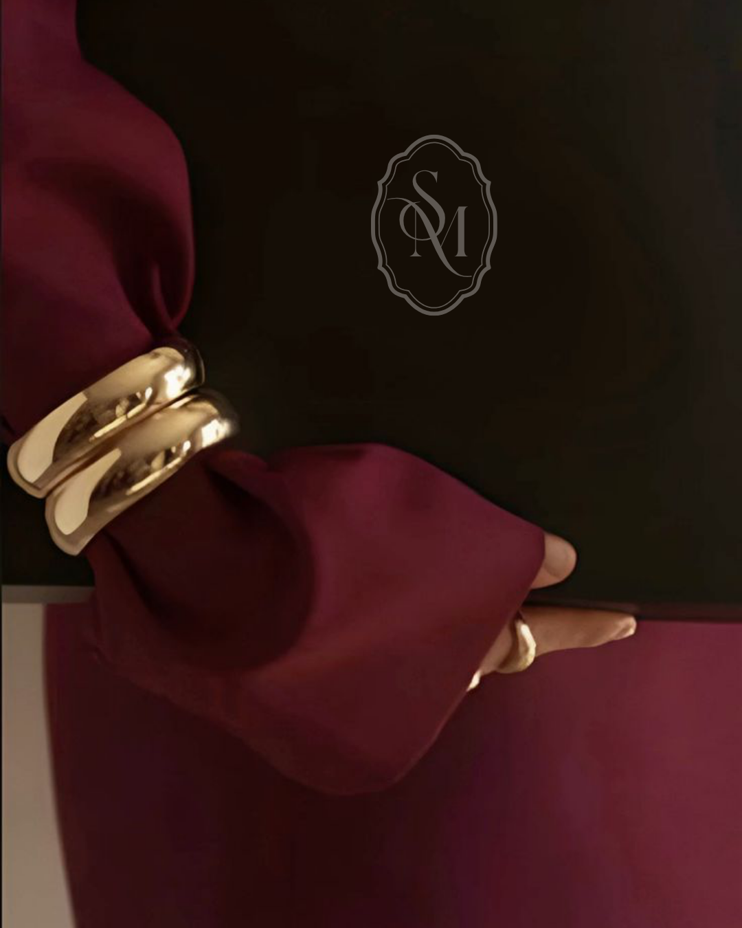 Close-up of a person wearing a black top, burgundy satin sleeves, and large gold bangles on their wrist, against a dark background with a decorative logo.