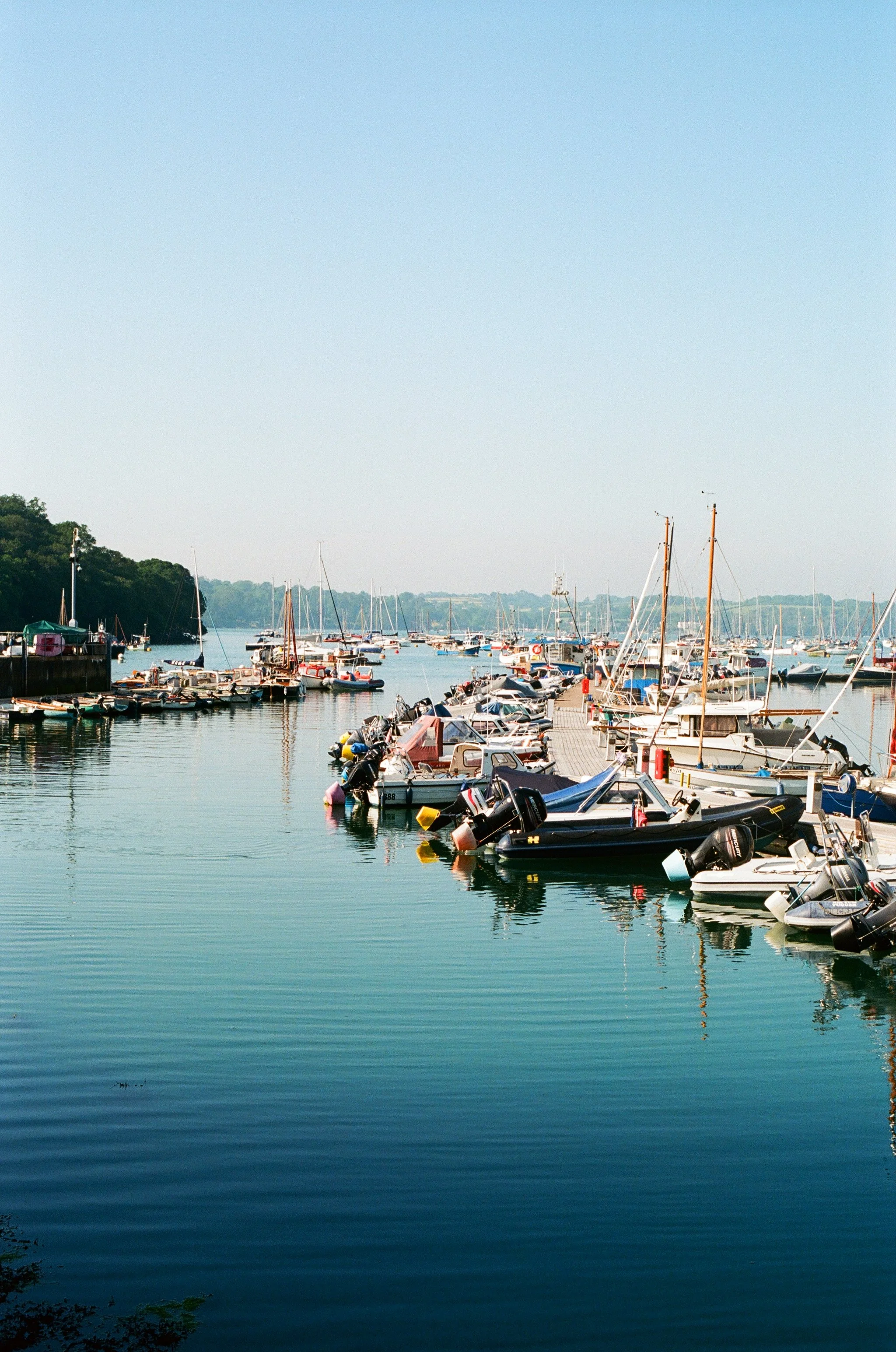 View of a marina filled with several boats docked along the piers, with a calm body of water reflecting the boats, and a distant tree-lined shoreline under a clear blue sky.
