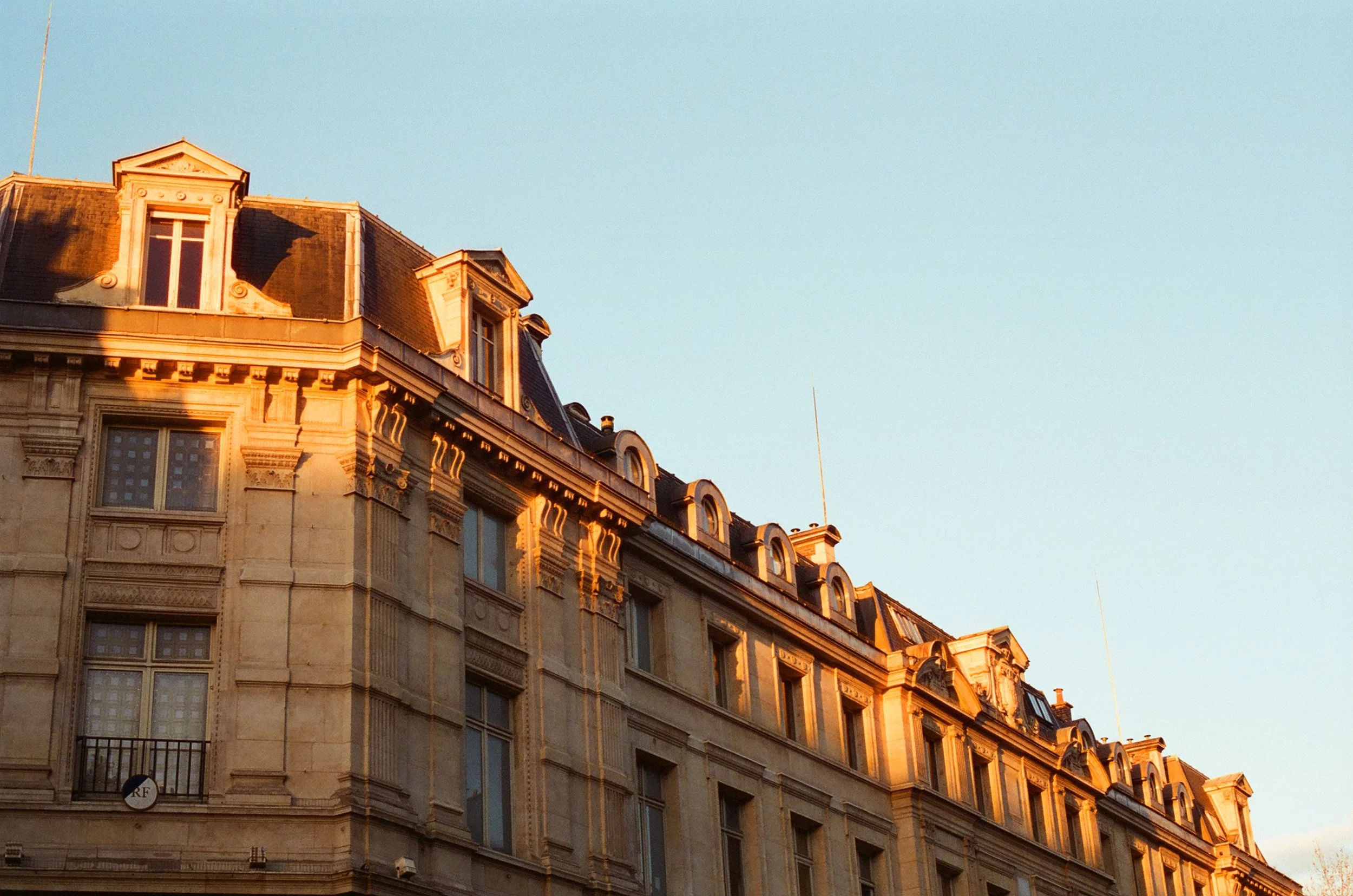 The upper story of a historic European building with ornate architecture, illuminated by warm sunlight, against a clear blue sky.