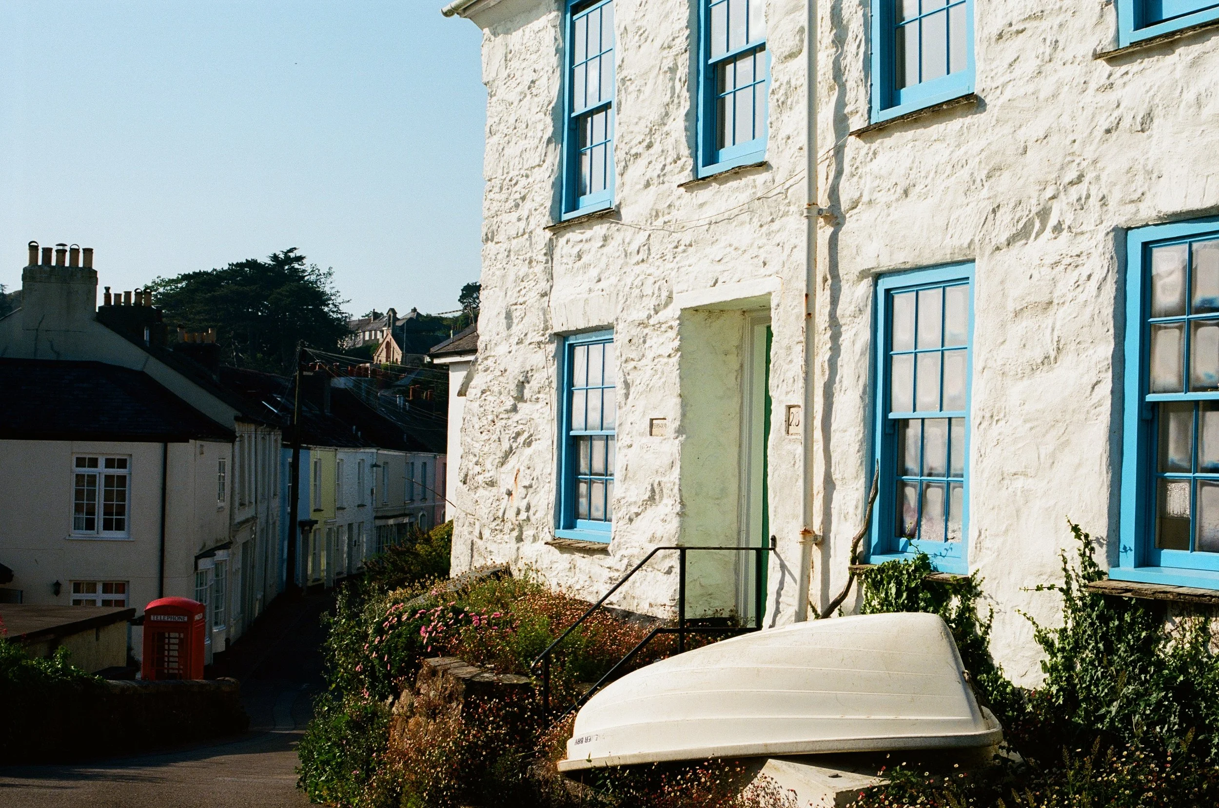A white, textured house with blue window frames and a blue door, situated on a hillside with houses in the background, greenery, flowers, and a boat in the foreground.