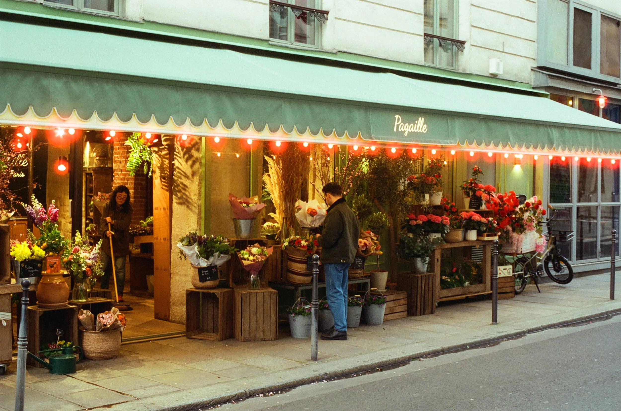 A flower shop called Pagaille with a green awning, displaying various colorful flowers outside. There is a man shopping for flowers and a woman working inside.