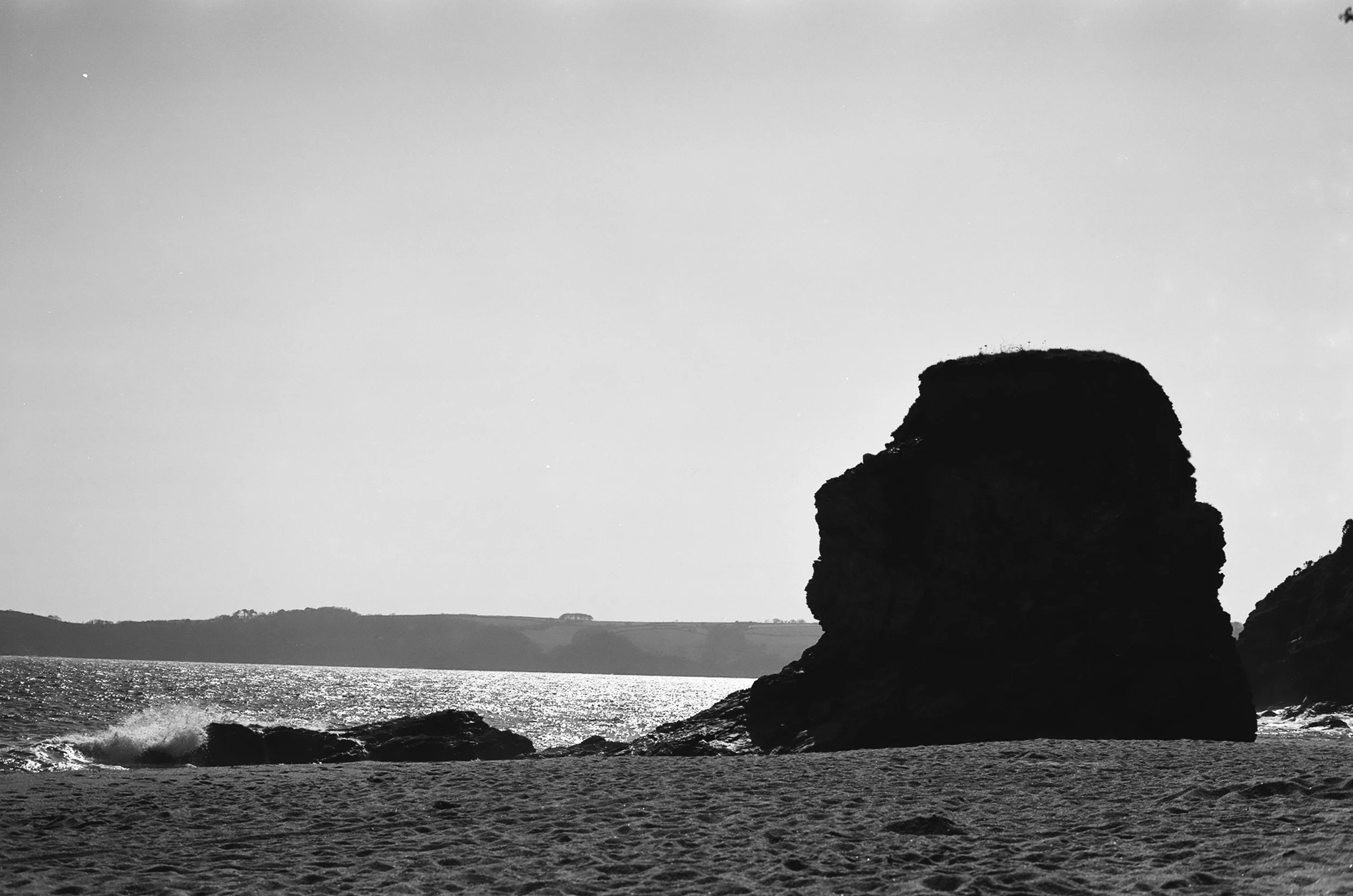 Black and white photo of a large sea stack rock formation on a sandy beach with water and distant hills in the background.
