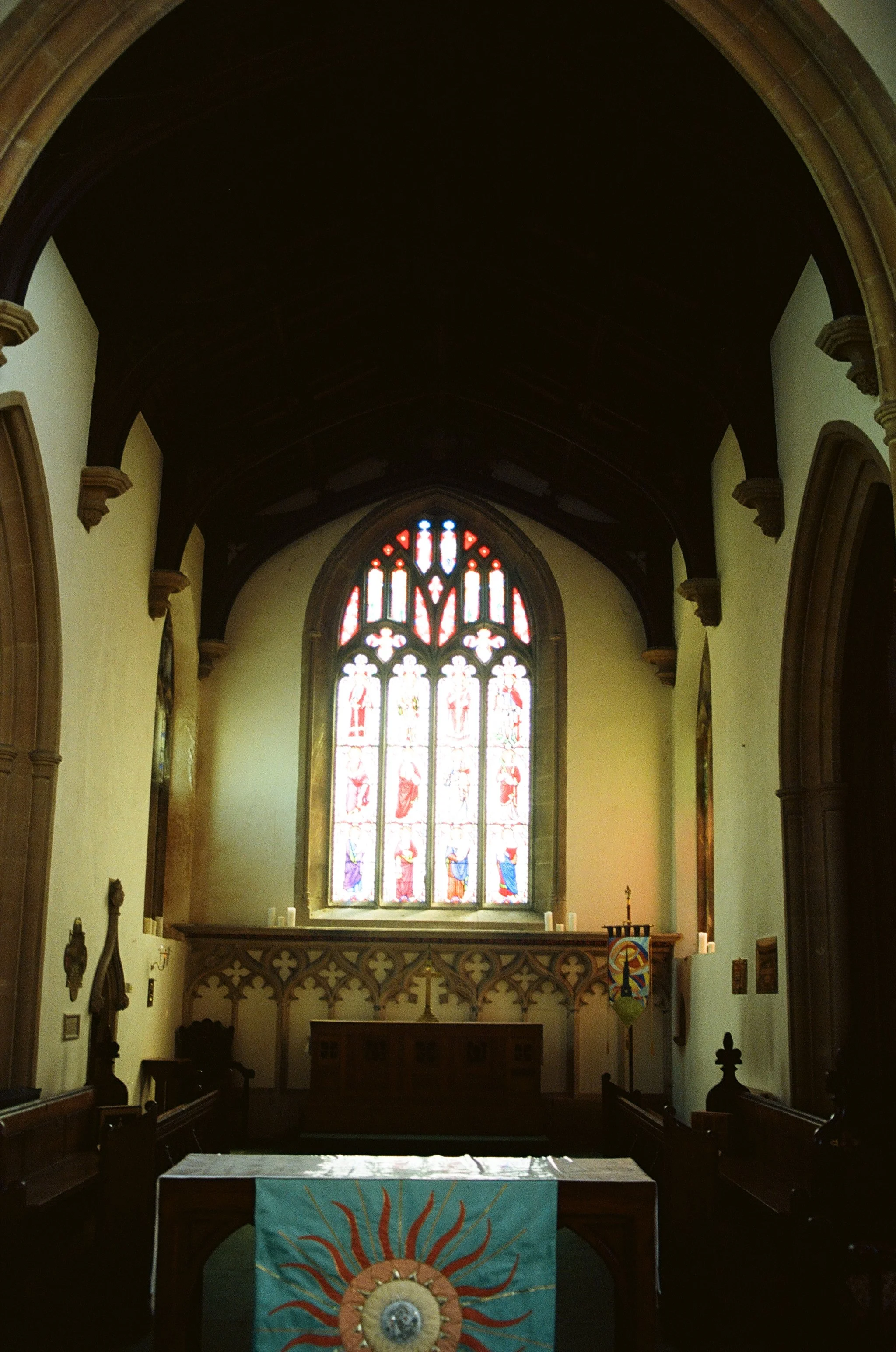 Interior of a church with stained glass window at the front and pews on either side. The altar has a colorful fabric with a sun and moon design.