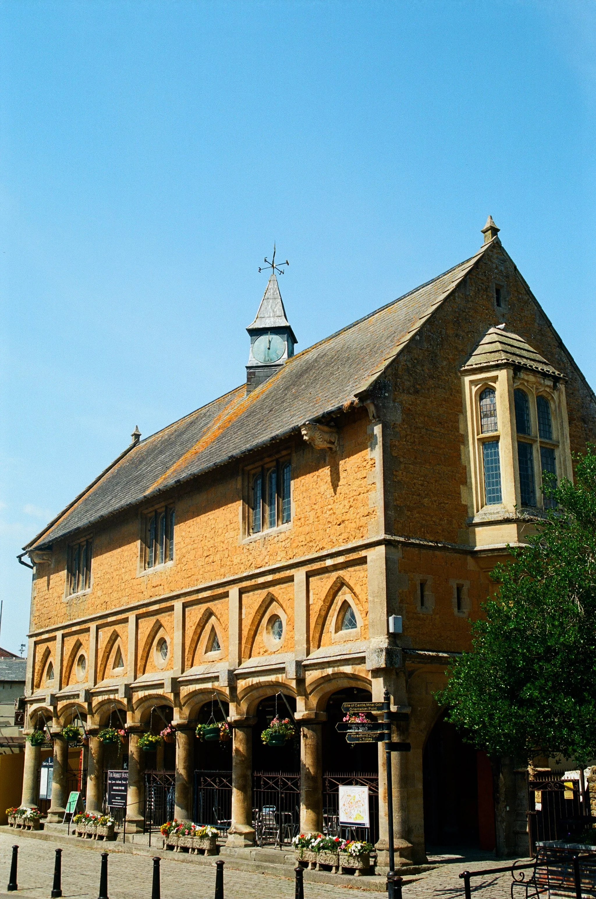 A historic stone building with arched and bay windows, a small clock tower with a weather vane, and a series of columns supporting a covered walkway. Flowers and street signs are visible in front of the building.