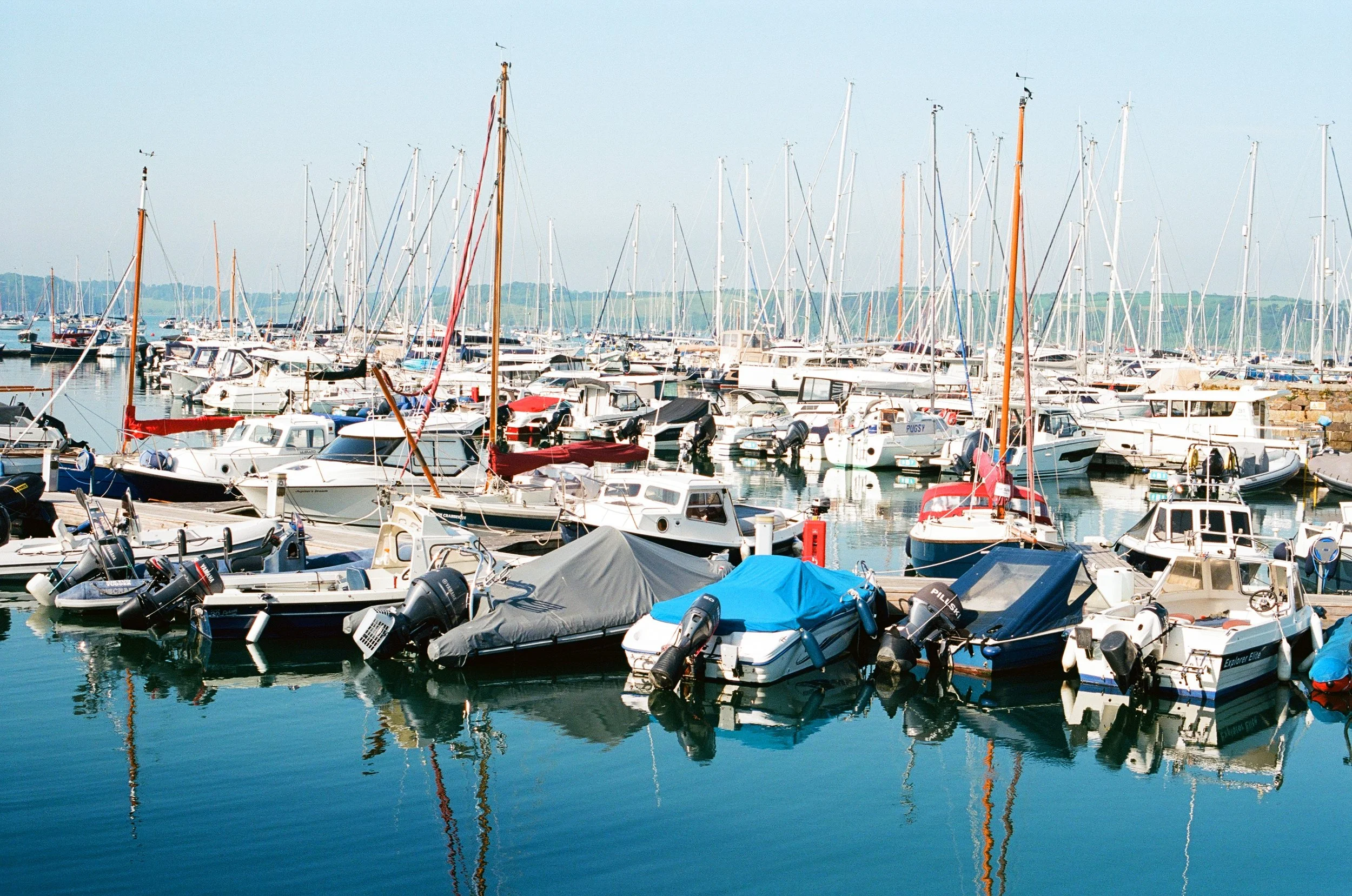 A marina filled with various boats and yachts docked in the water, with masts visible and a distant shoreline with greenery.