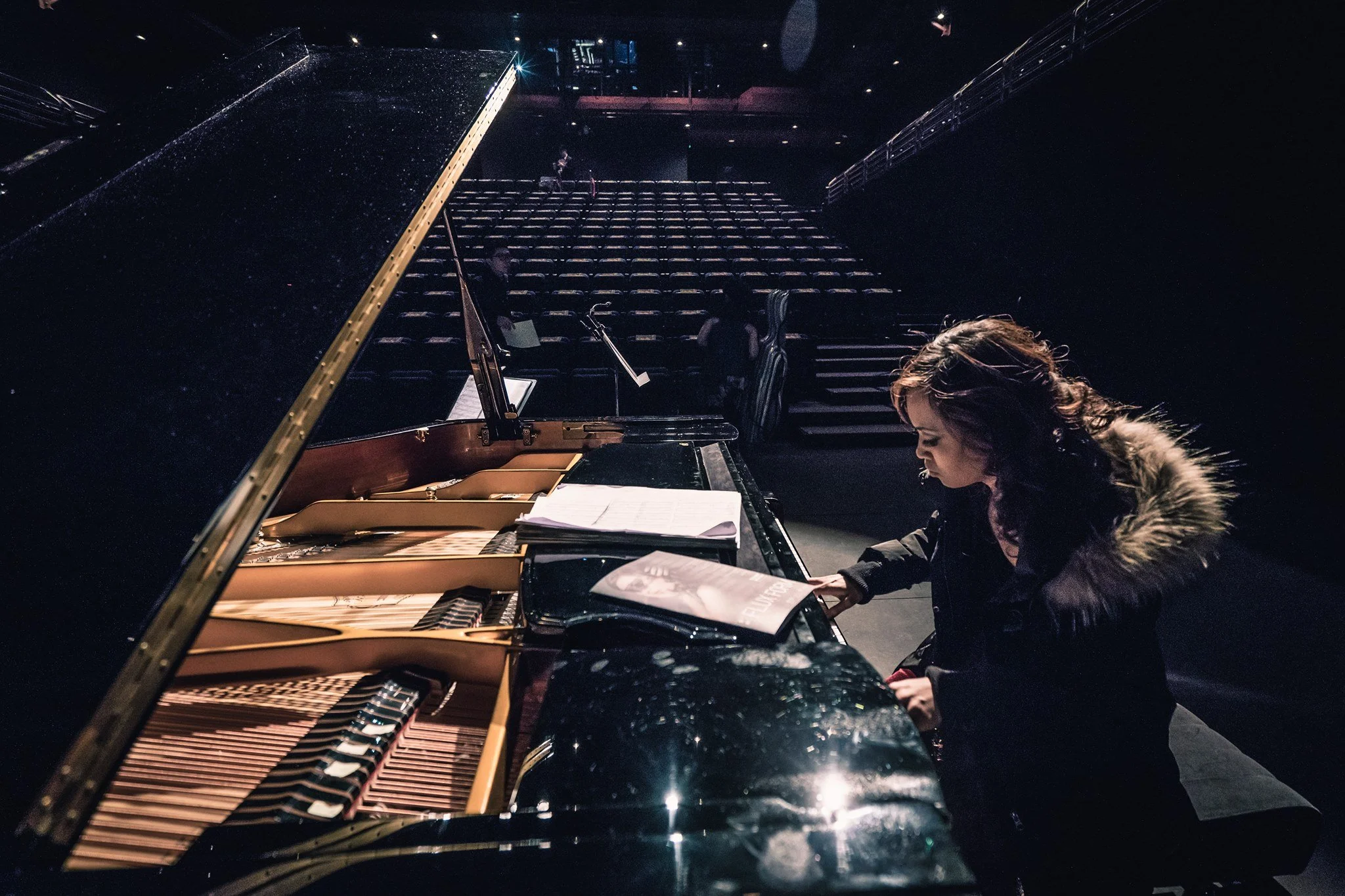 A woman with long brown hair wearing a black coat with a fur collar playing a grand piano on stage in a dimly lit theater with empty seats.