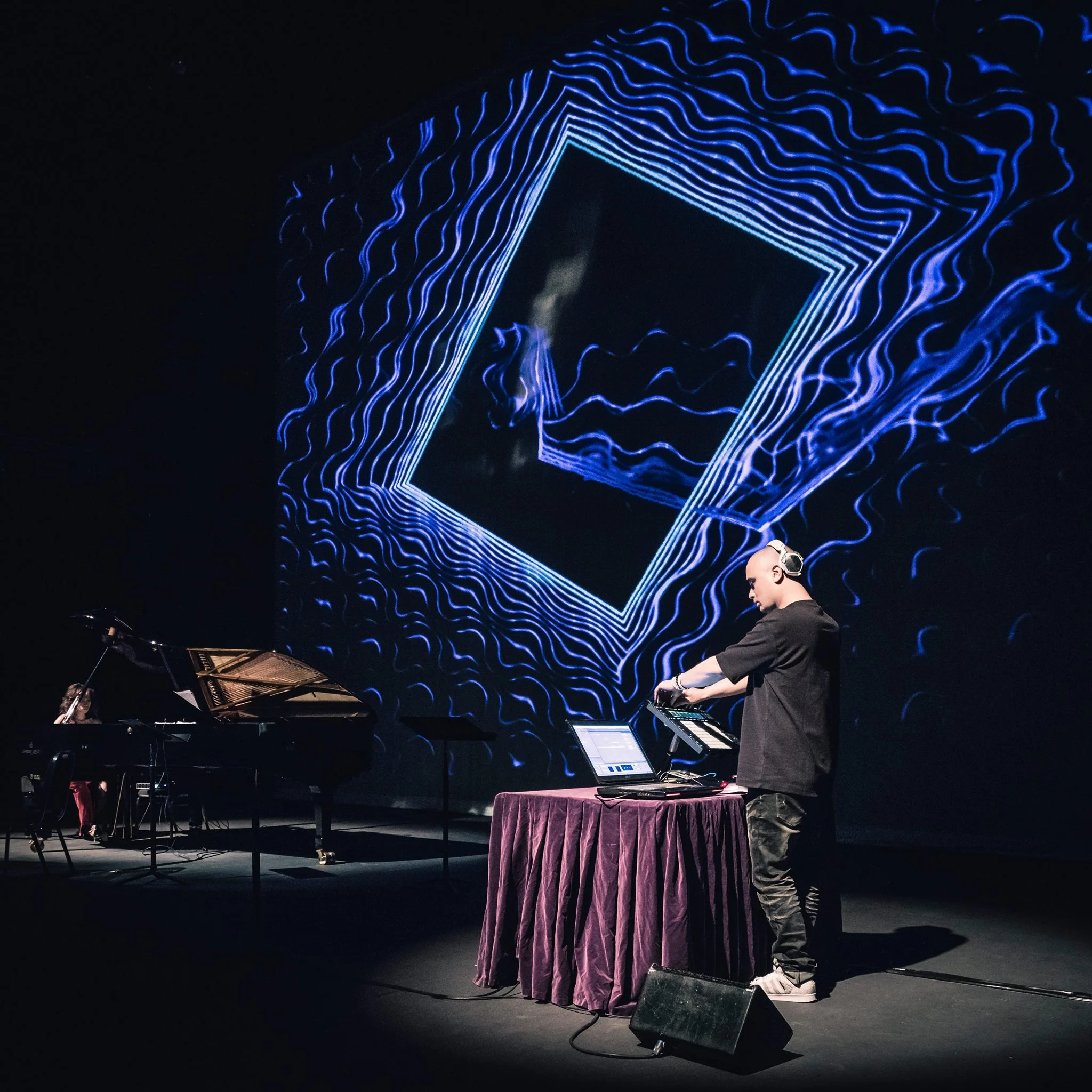 A person in casual clothing and sneakers is performing on a stage in front of a large dark screen with glowing blue geometric and wavy line patterns. To the left, a pianist is playing a grand piano. A table with a purple tablecloth and some electroni