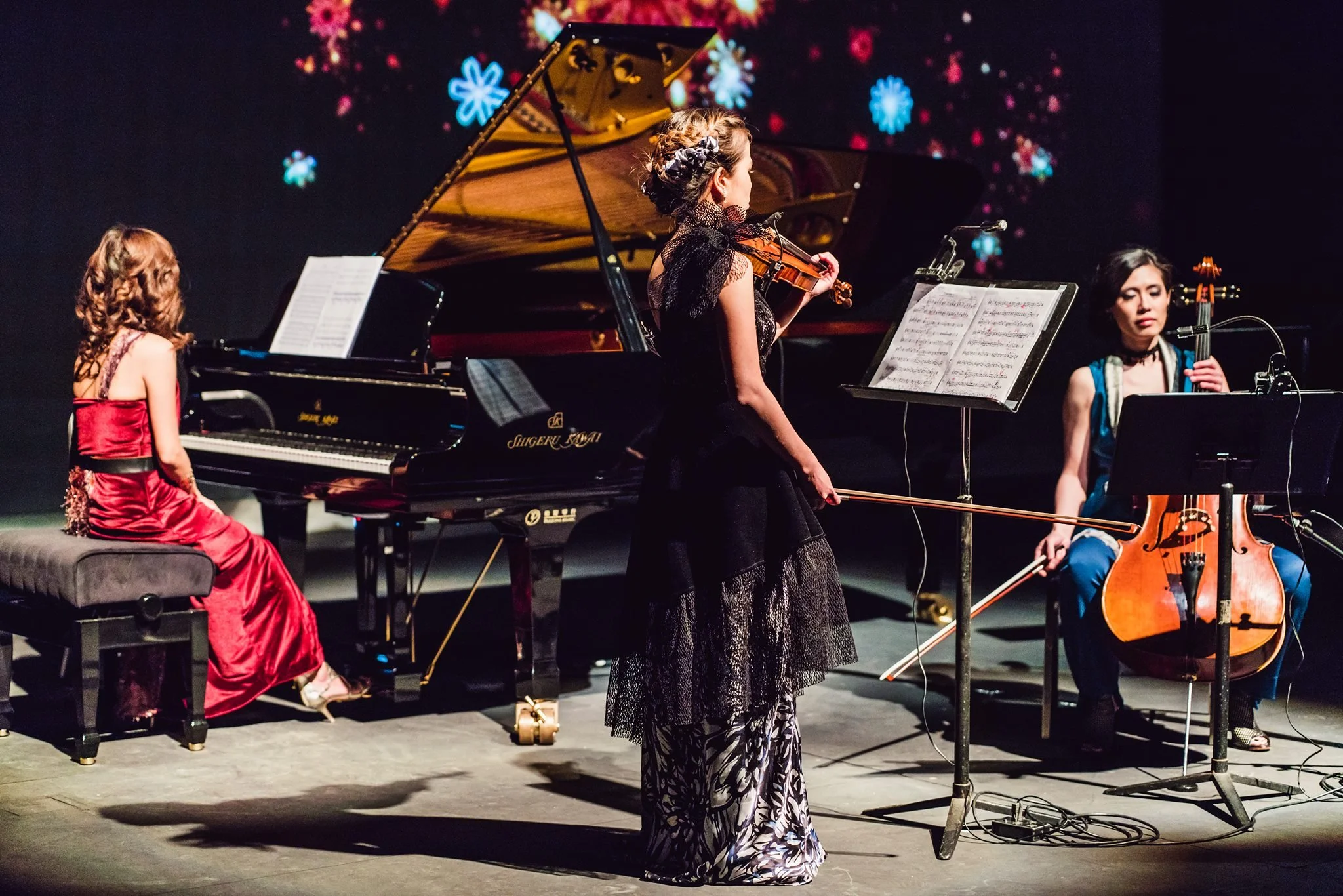 Three women performing on stage with a grand piano, a cello, and music stands with sheet music, behind a dark backdrop with colorful flower patterns.